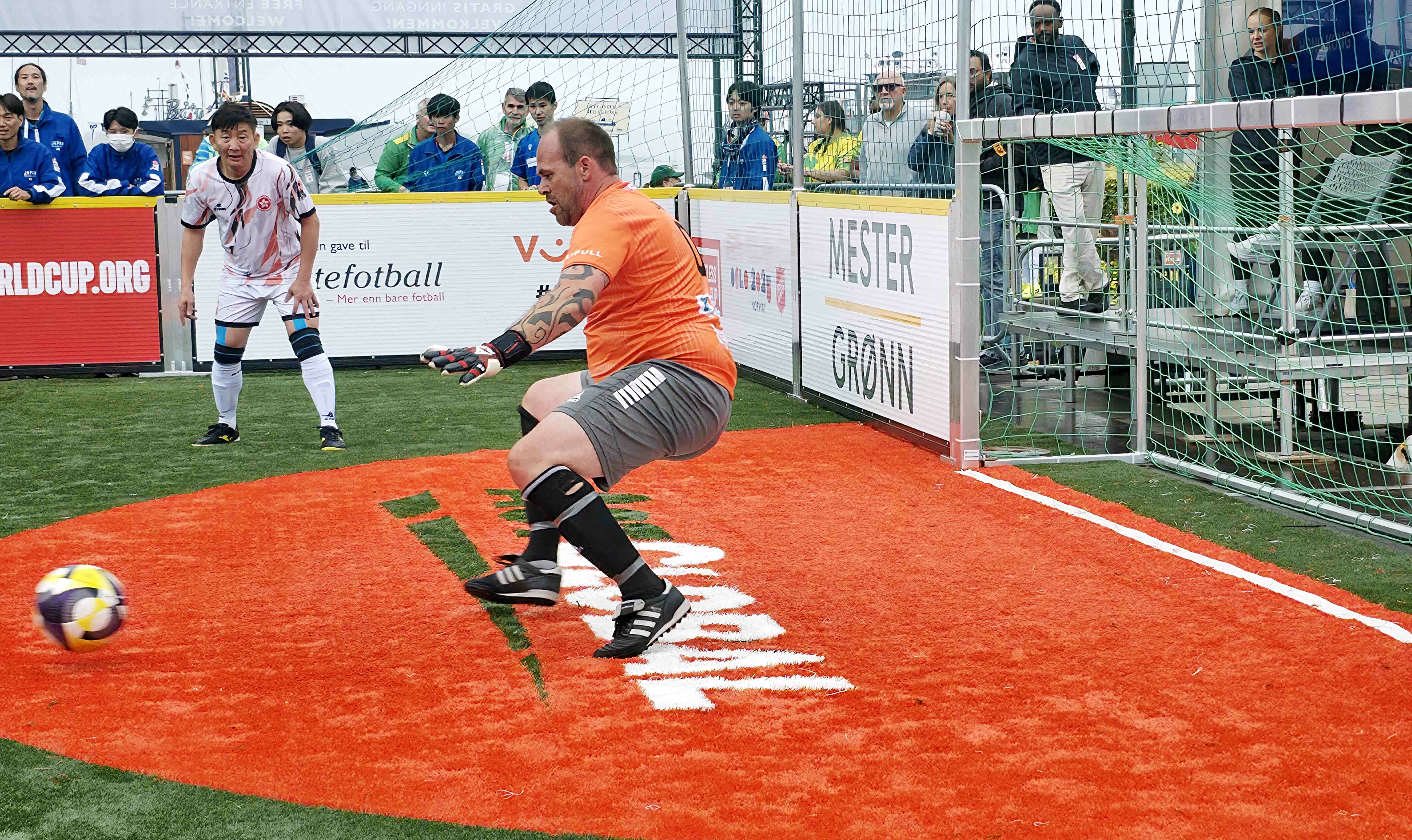 Foto: Bilde fra an kamp under VM. En keeper i oransje t-skjorte og grå shorts har akkurat sparket ballen bort fra mål. Ballen sees i kanten av bildet. En spiller i hvit drakt står litt framoverbøyd og følger med.