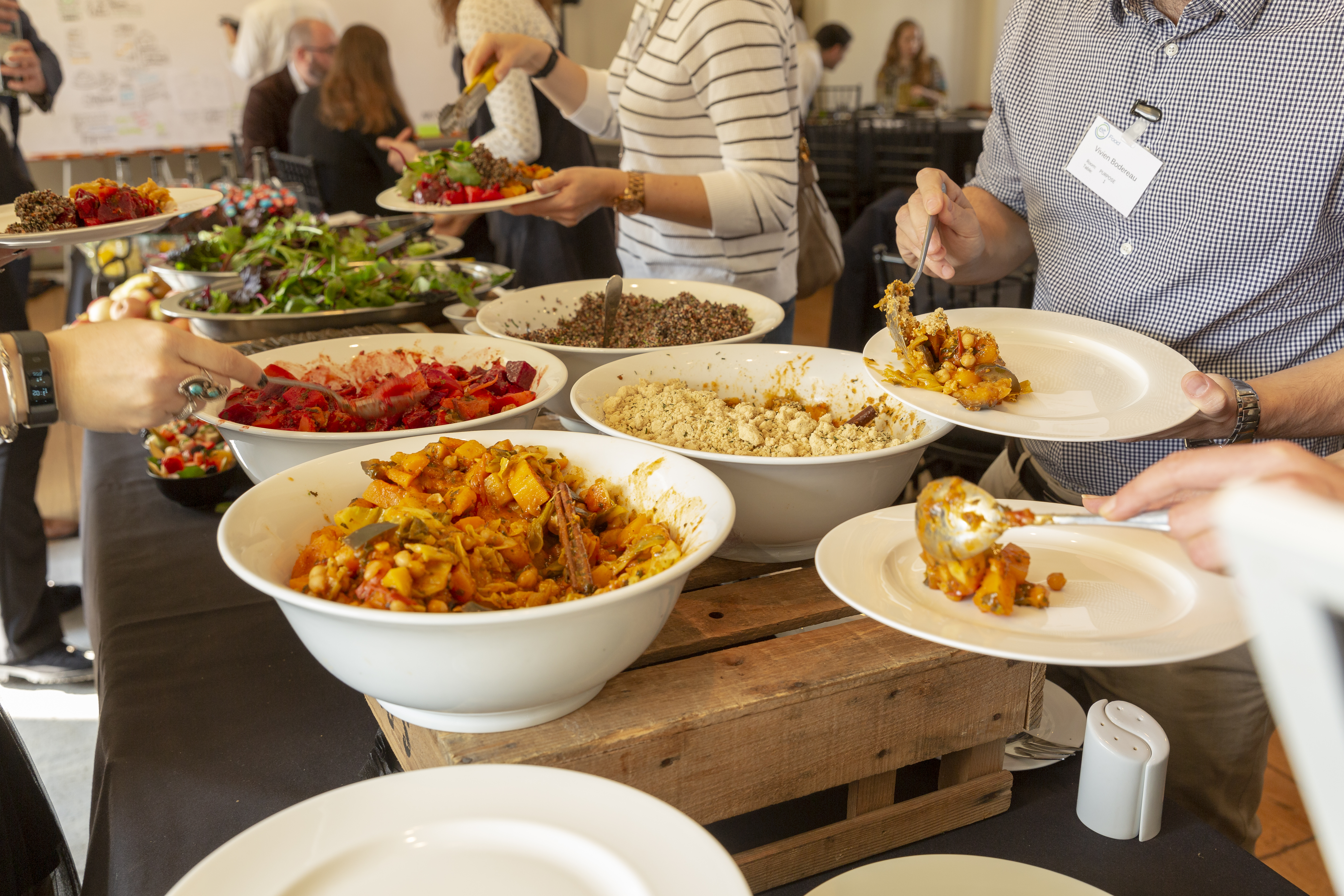 A selection of food in white bowls is placed on a wooden table. The food is different colours and a selection of people help themselves.