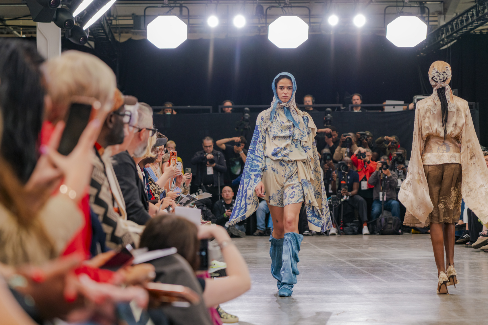 Models walking down runway in flowing blue and beige garment, people blurred watching in front.