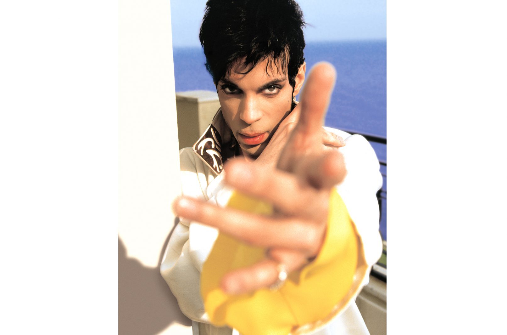 Prince in a white shirt extending a hand towards camera, sea in the background.
