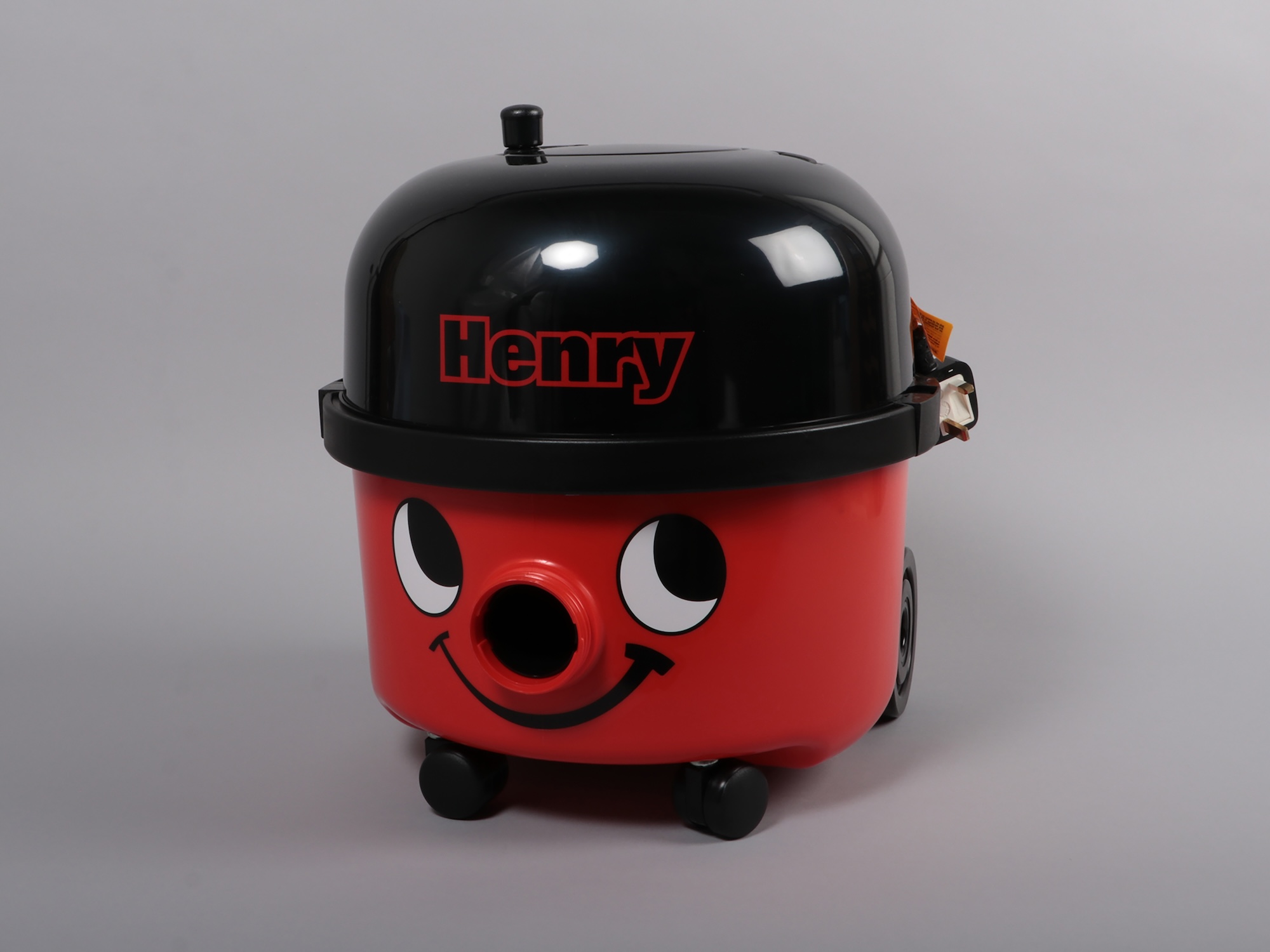 A red Henry vacuum cleaner with a black lid and smiling face sits on a plain grey background.