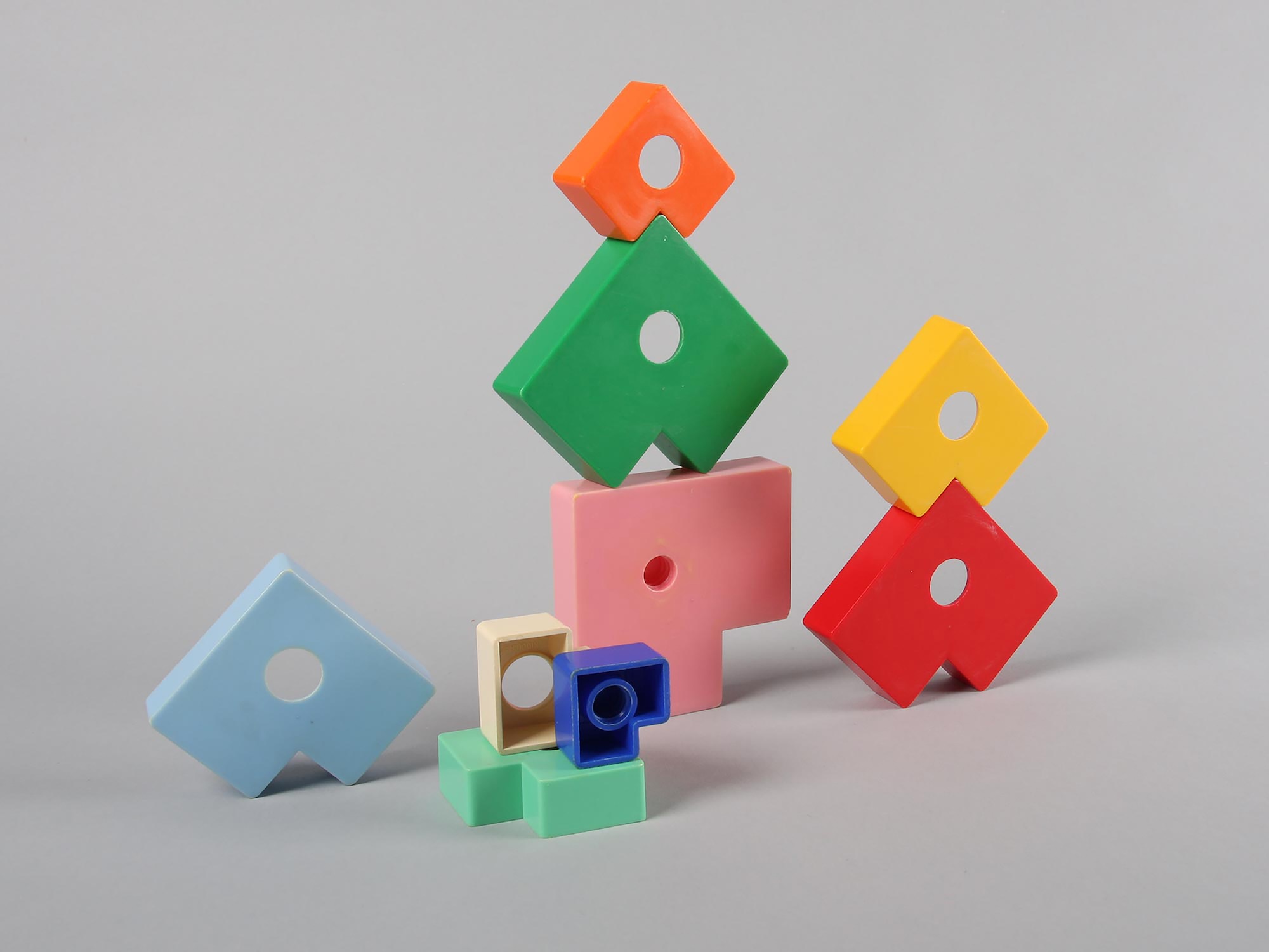 Colourful geometric toy blocks stacked on a grey surface, including a green diamond-shaped piece topped with an orange block and a red and yellow pair to the right.