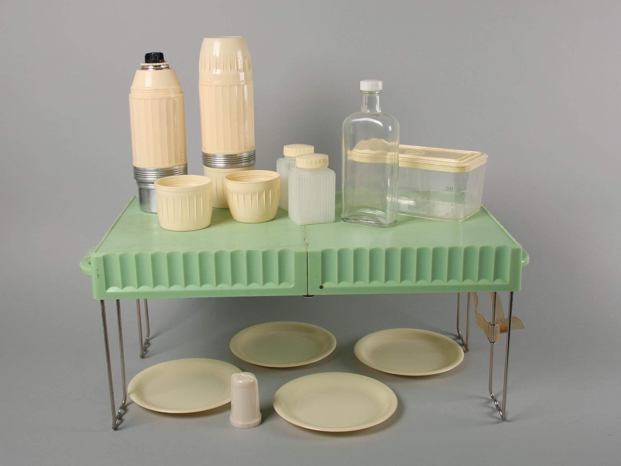 Various cream and clear glass containers arranged on mint green plastic rack with metal legs, cream plates beneath on grey surface.