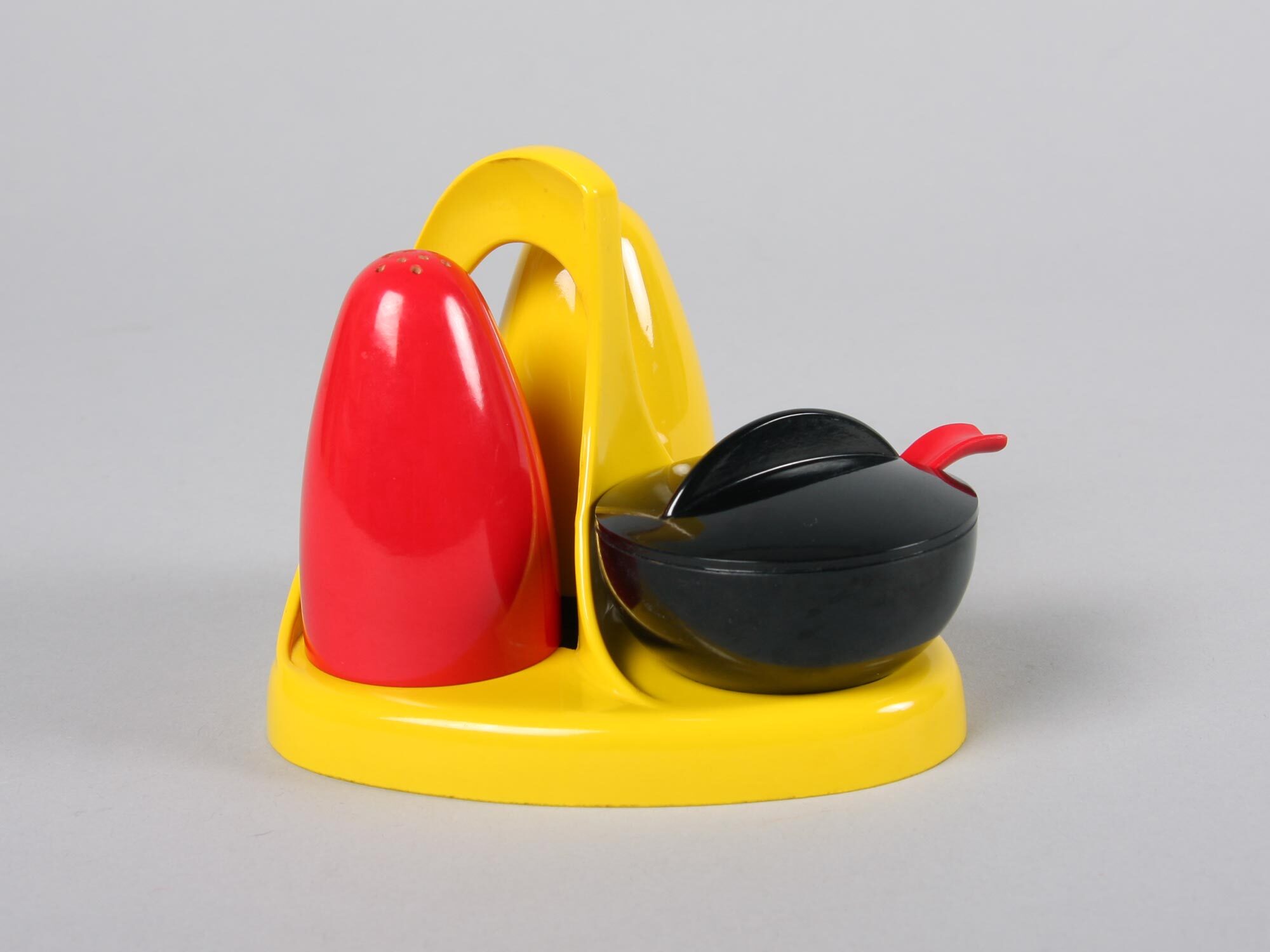 Three-piece salt and pepper set on yellow plastic base with red dome-shaped shaker and black ovoid container with red spout on grey surface.