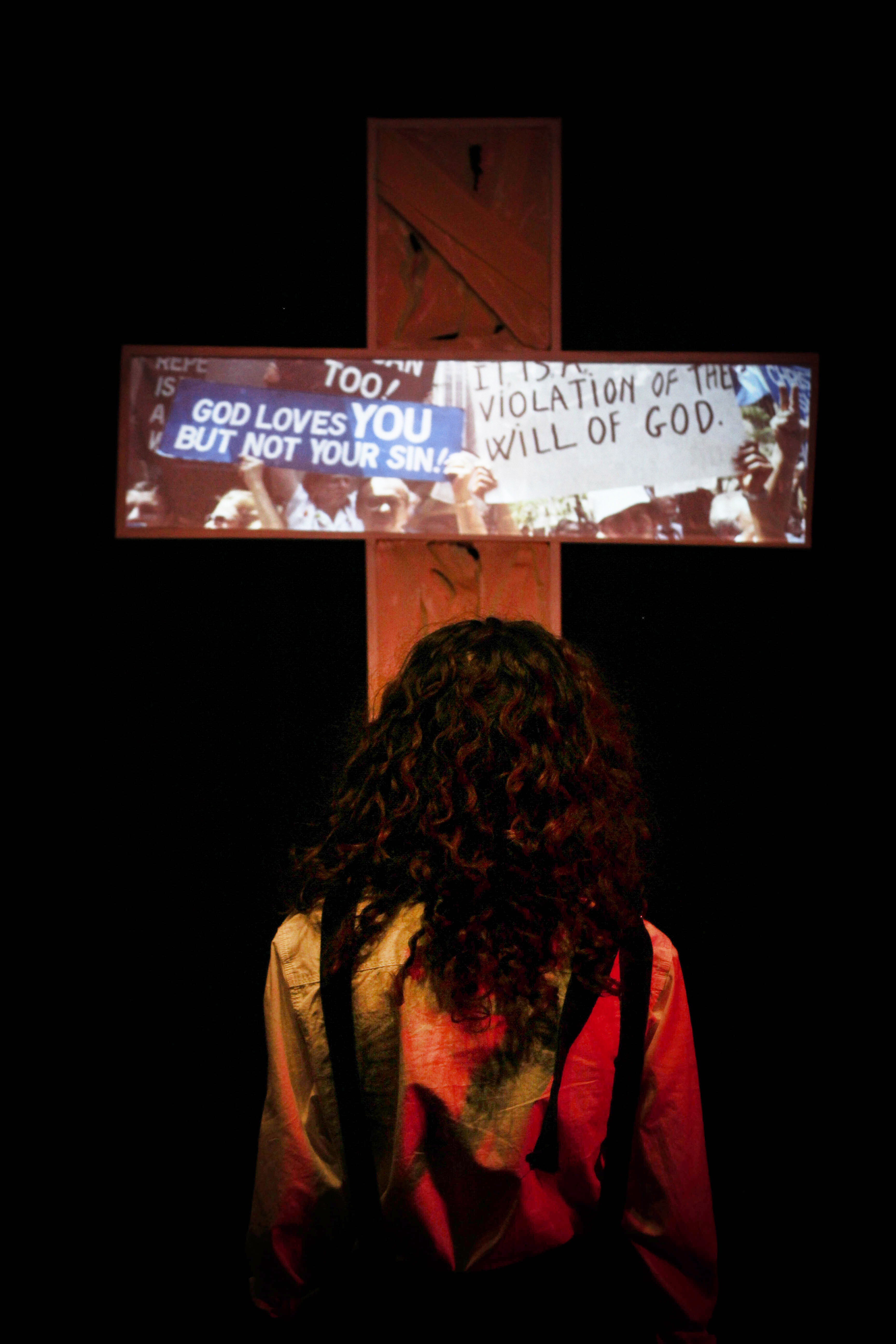 An actor with long, curly hair sits in front of a cross with a projected image from historic LGBT protests, dark background.
