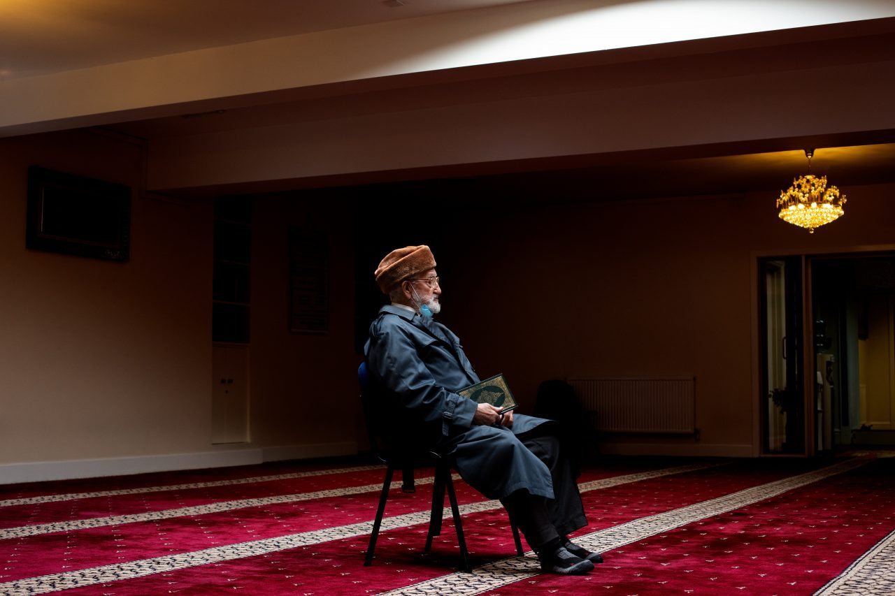 An old man sits alone on a chair in a large empty room.
