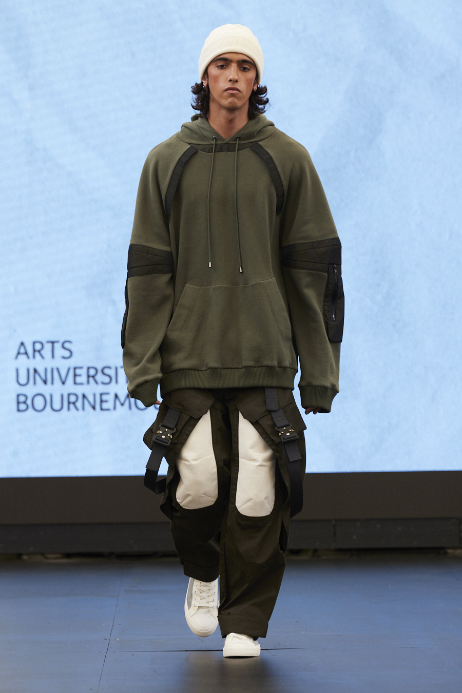A model wearing a white hat and dark olive green hoody walks across a catwalk
