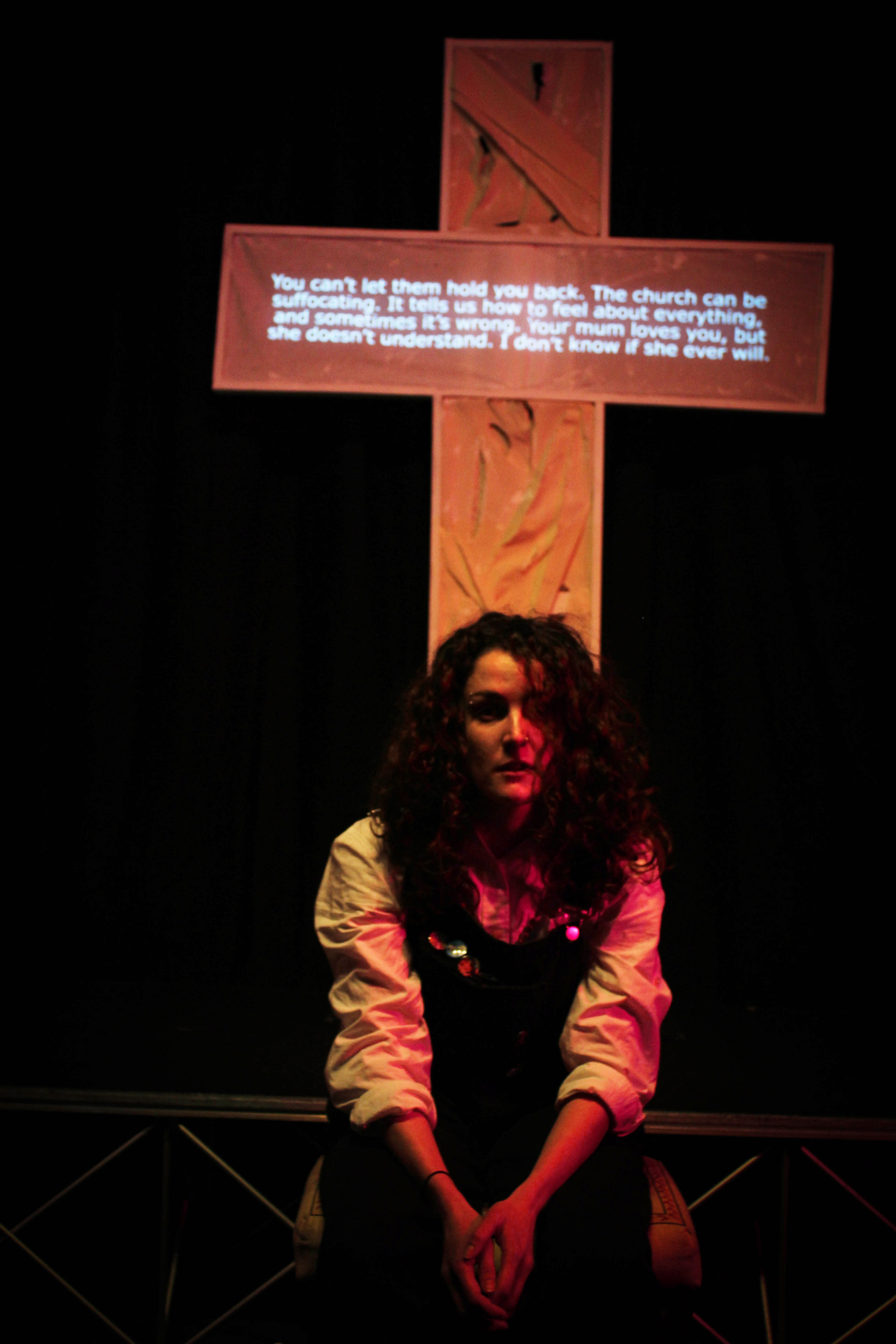 An actor with long, curly hair sits in front of a cross with projected text, dark background.