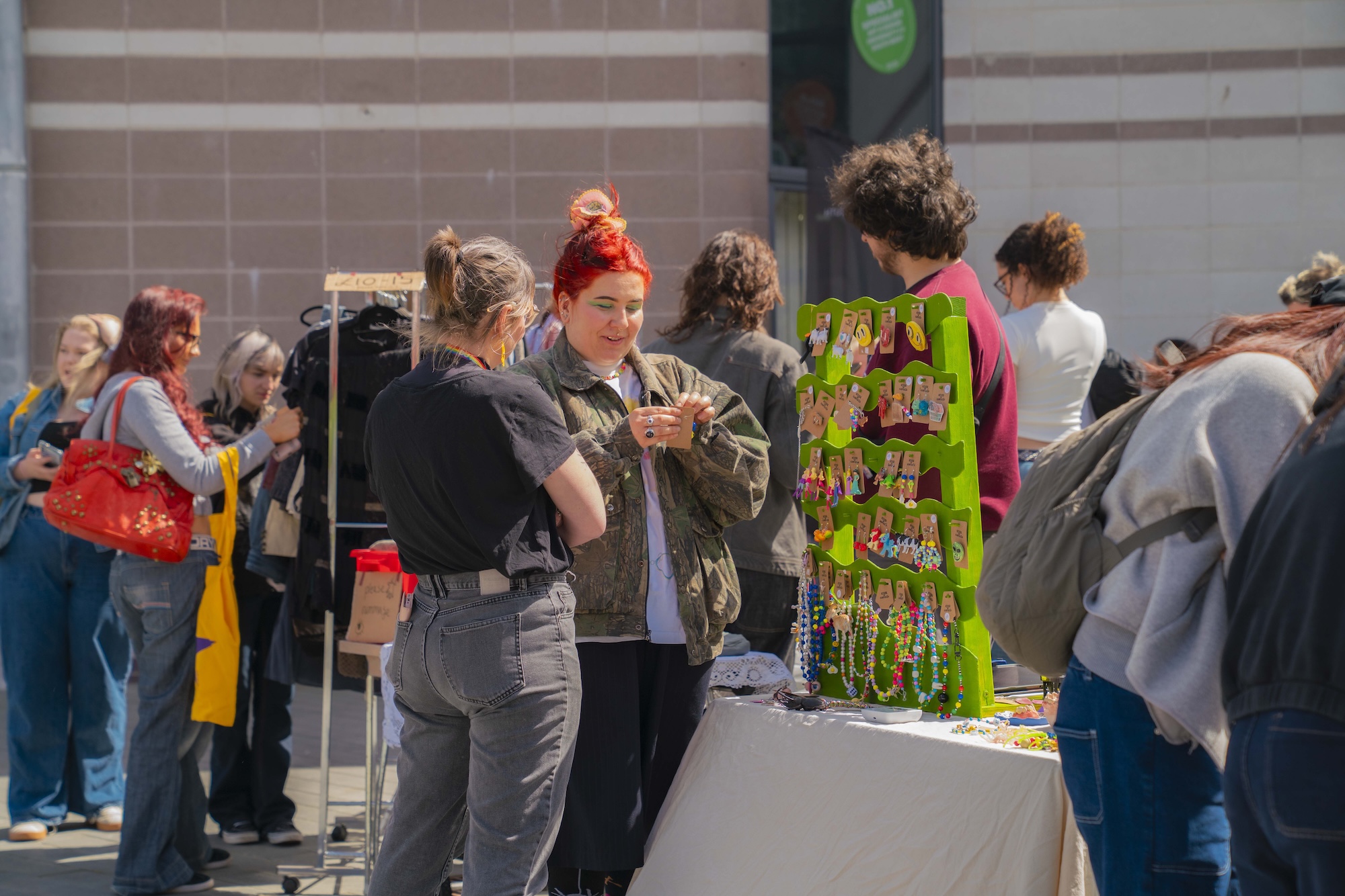 A group of people browse a stall with colourful hanging jewellery and trinkets at an outdoor market beside a brick building.