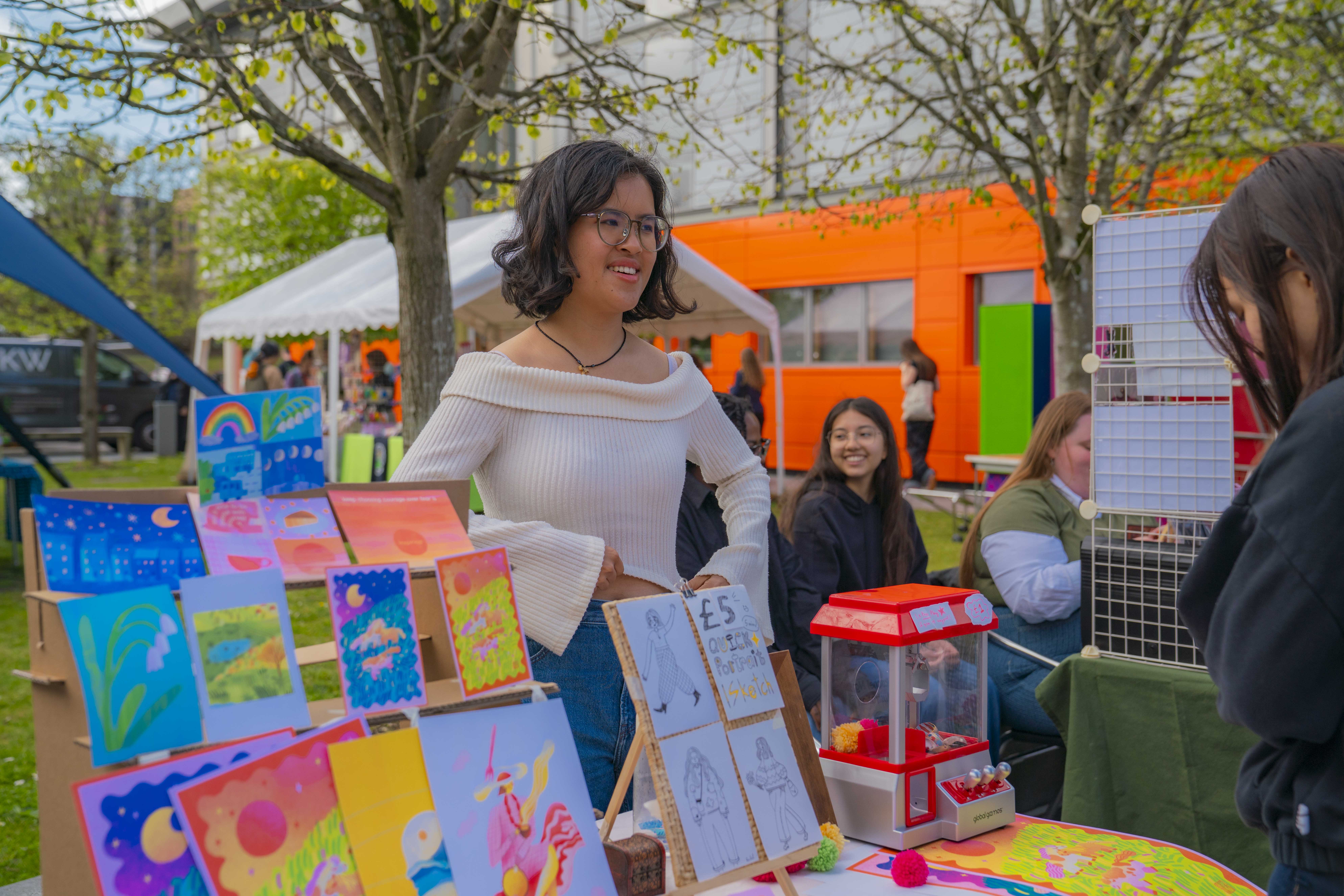 Student stood smiling at a stall with bright, colourful illustrations in AUB courtyard, trees and orange South House building in the background.