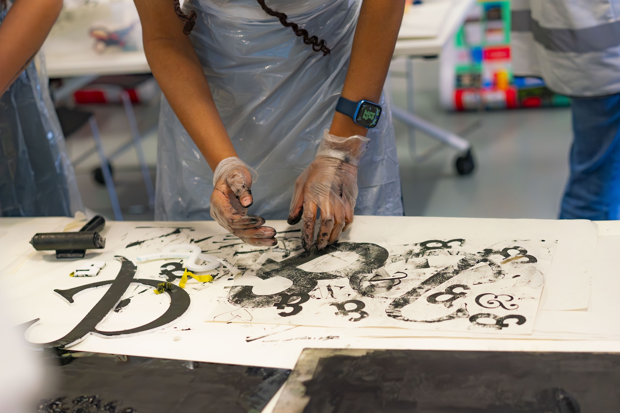 A person wearing gloves uses ink-covered hands to print abstract shapes on a large white sheet at a work table, with art supplies and a blurred studio background.