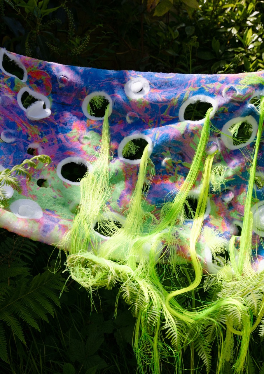 Textured textile fabric with holes in and green hair-like textures, suspended in a forest.