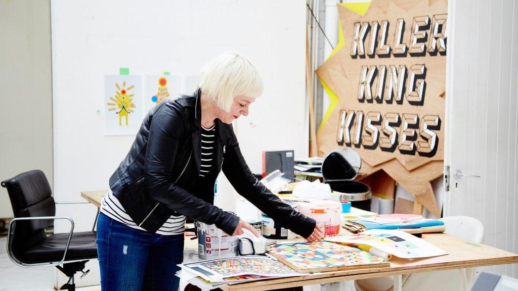 A person stands over a table with various craft items on it. A board in the background reads Killer King Kisses