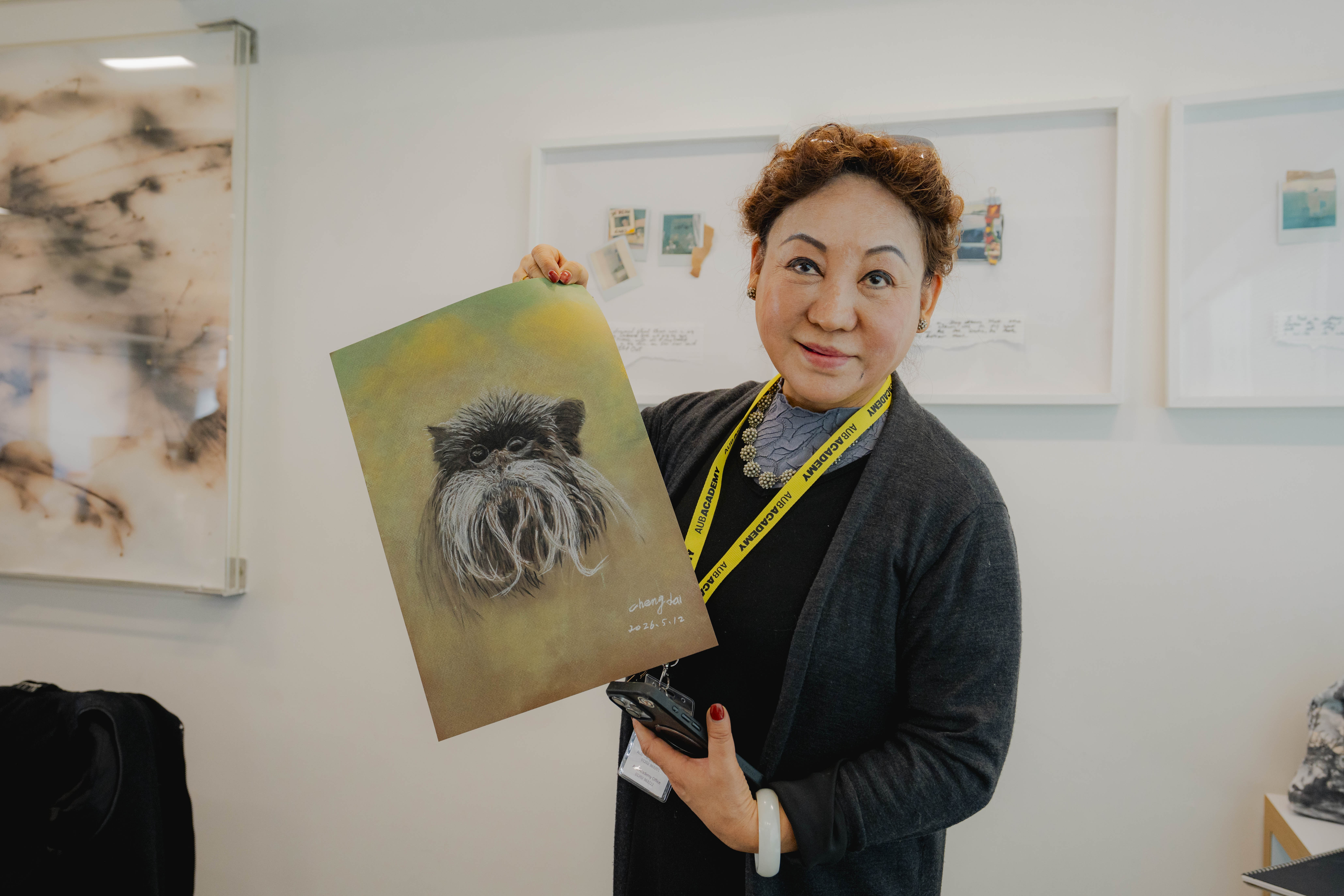 A person holding up a framed artwork of a shaggy dog portrait, standing in a gallery with mounted pictures on the white wall behind her.