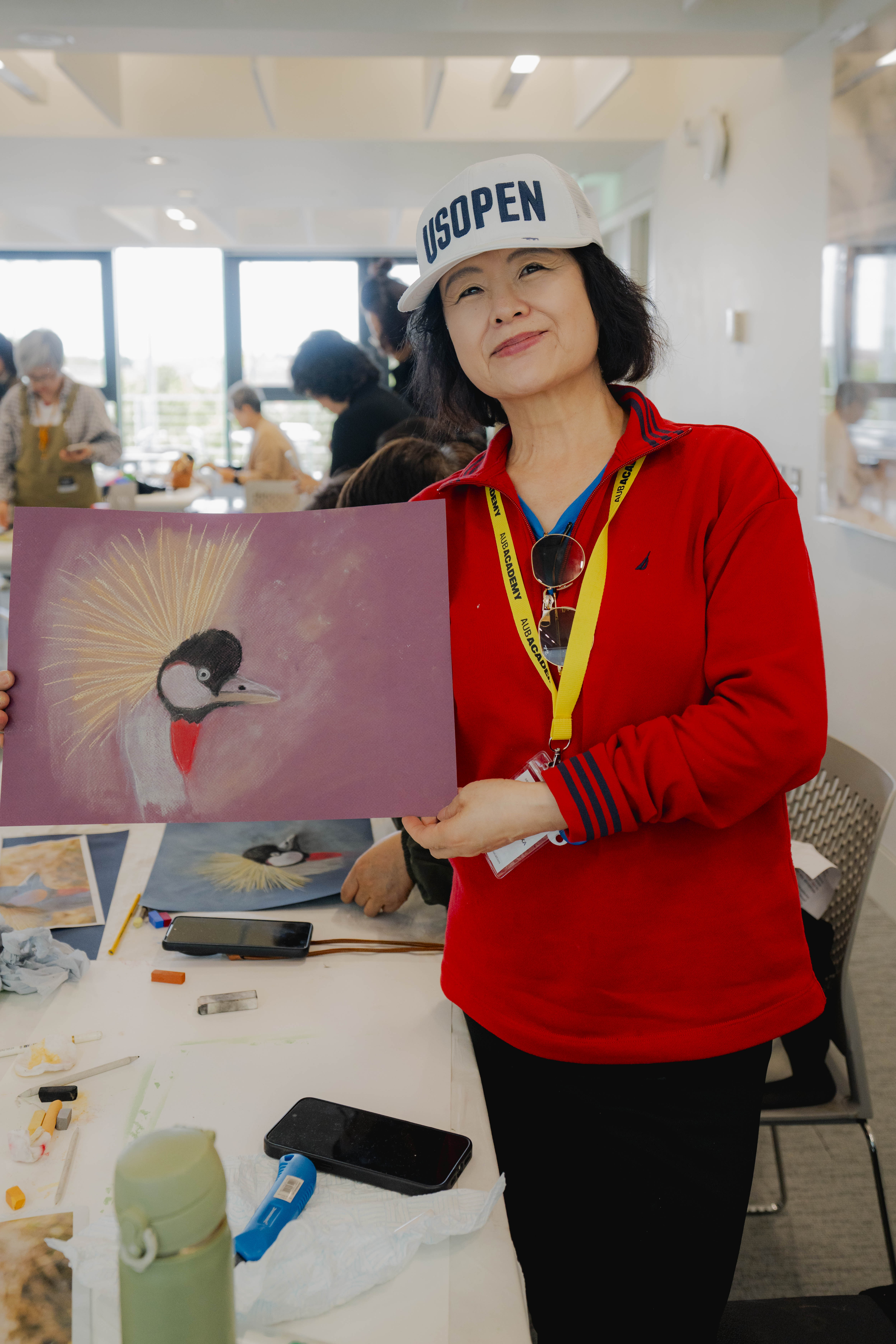 A person in a red top and white cap stands indoors holding a painting of a bird, with people seated at a table and large windows in the background.