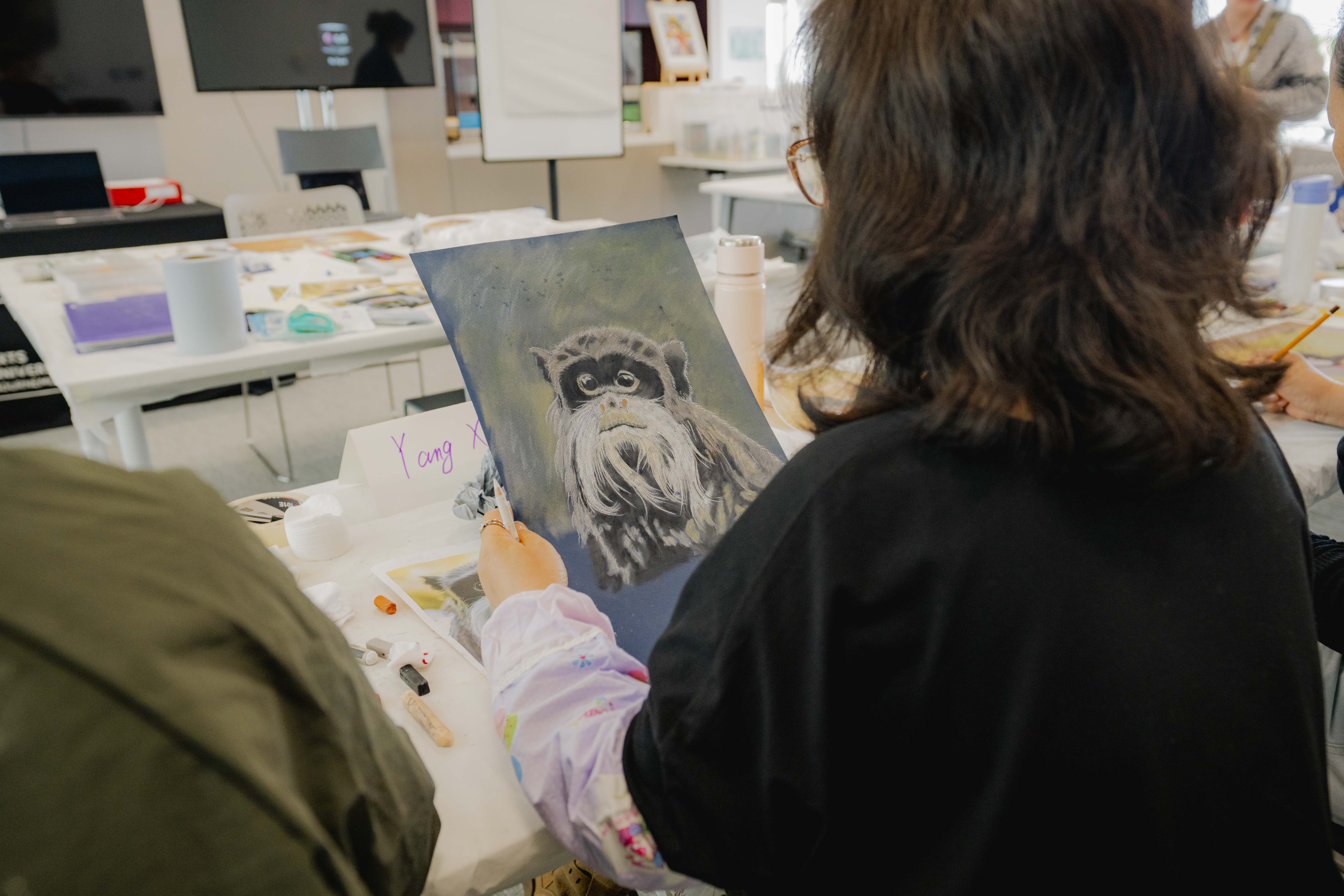 A person paints a sloth portrait on a canvas at a table, with art supplies and other participants visible in the background.