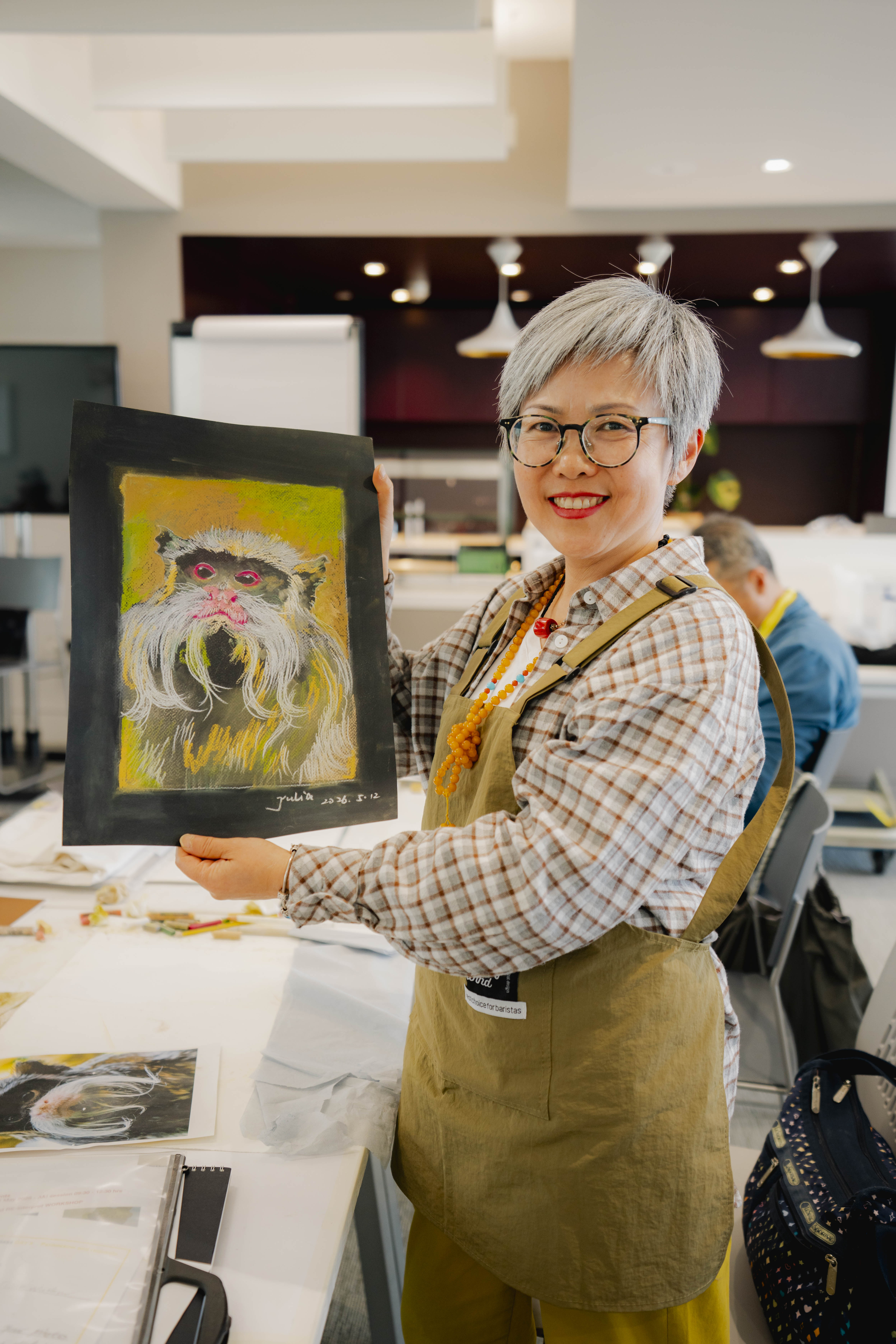 A person with short grey hair and glasses holds a framed dog painting in a bright indoor studio, with art supplies and another seated person in the background.