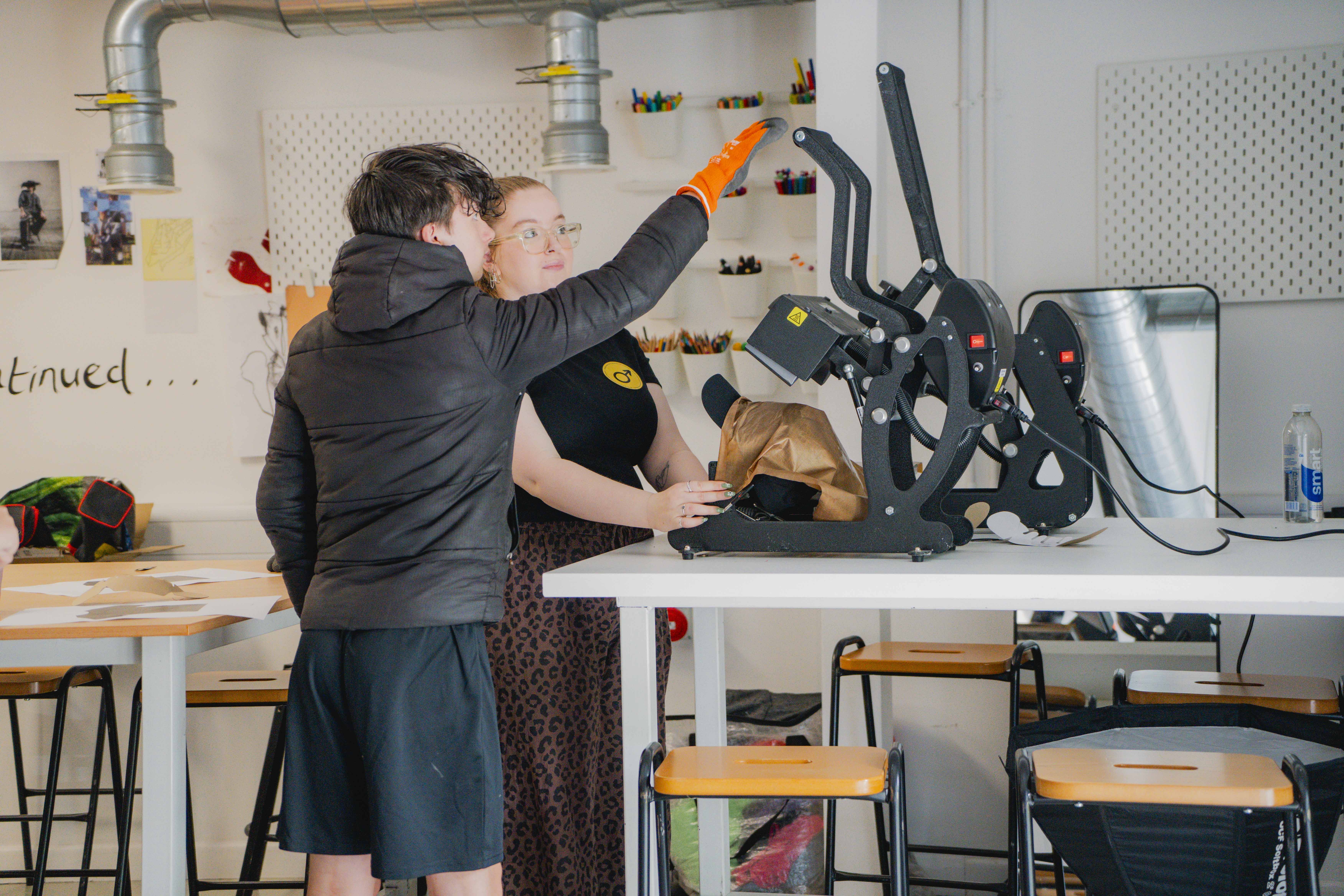 Student wearing a black coat and shorts with teacher operating equipment, orange glove and white workshop background.