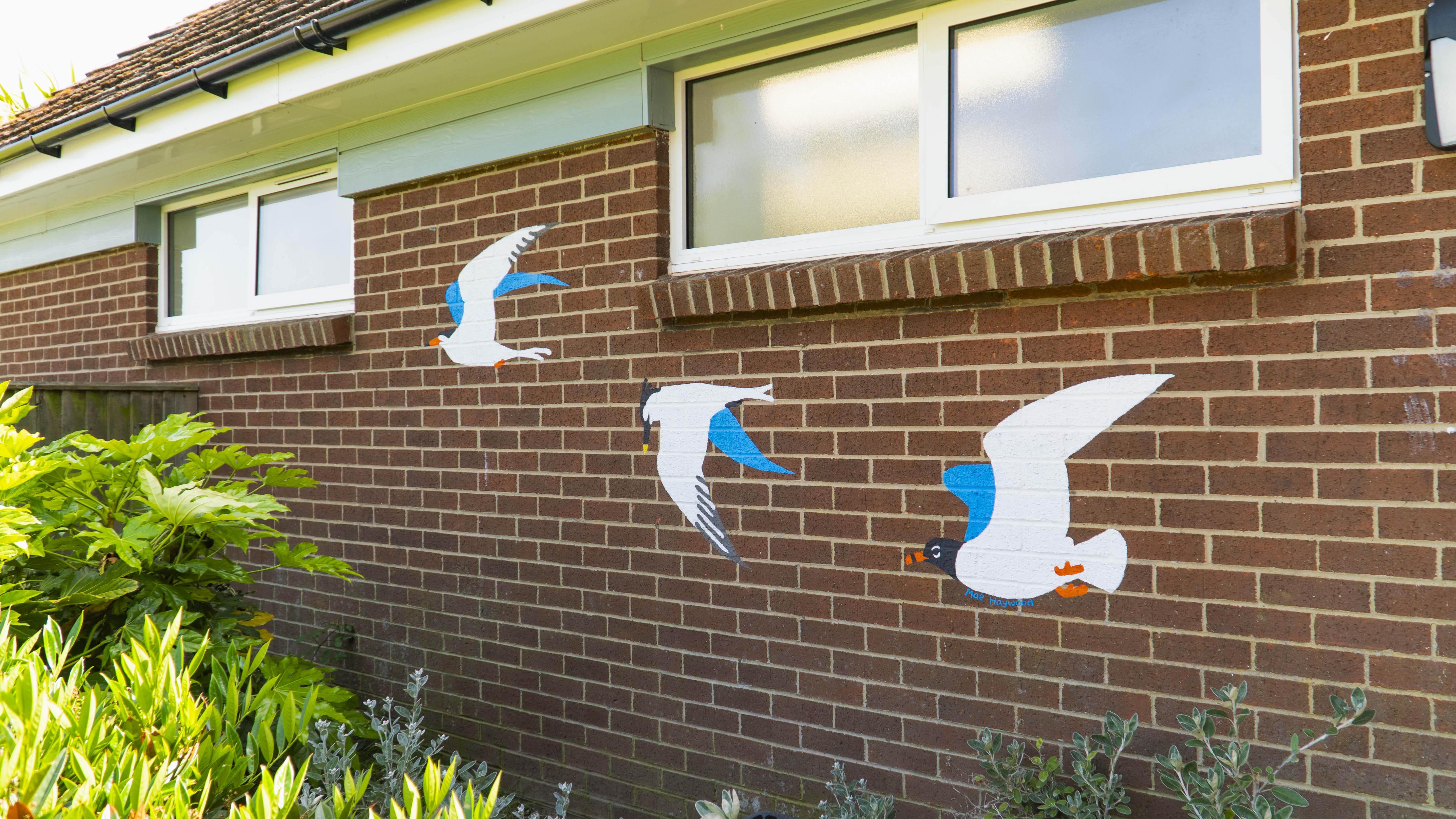 Brick wall of a building with a mural of three sea gulls under its windows.