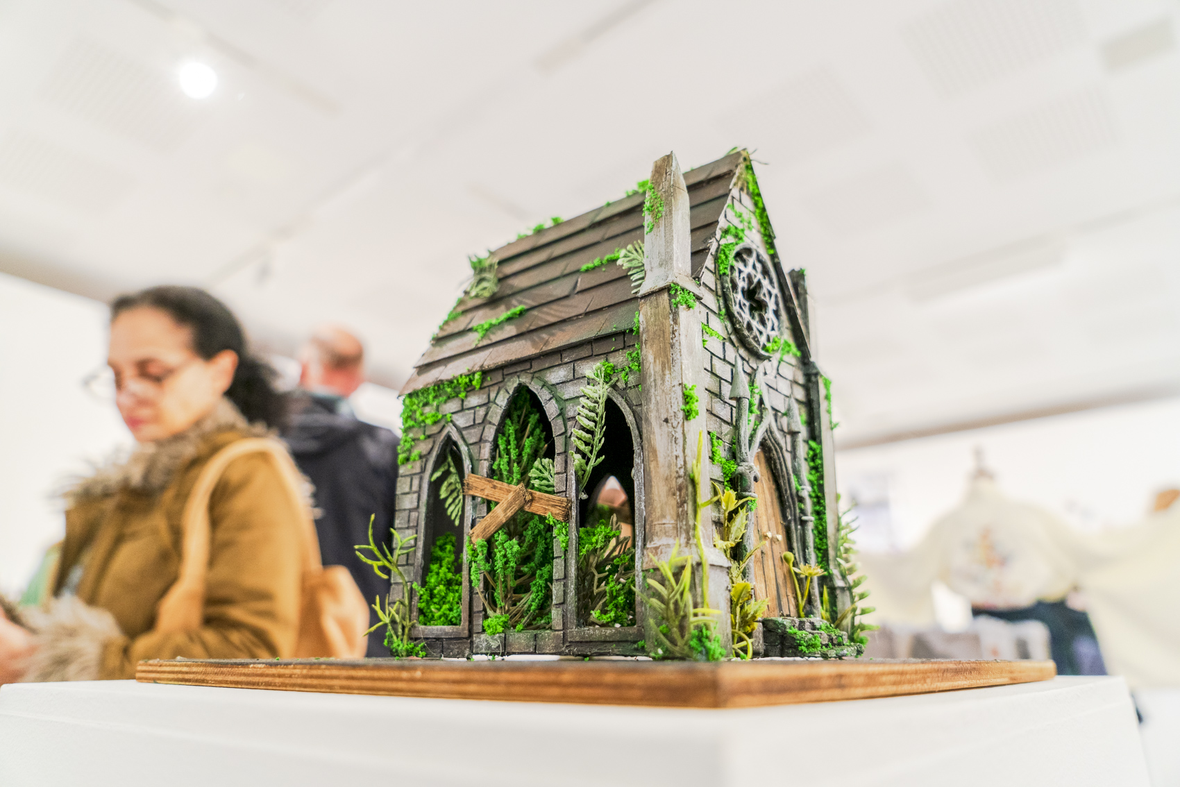 A detailed miniature stone chapel model with arched windows and green foliage sits on a wooden base, with blurred people in the background.