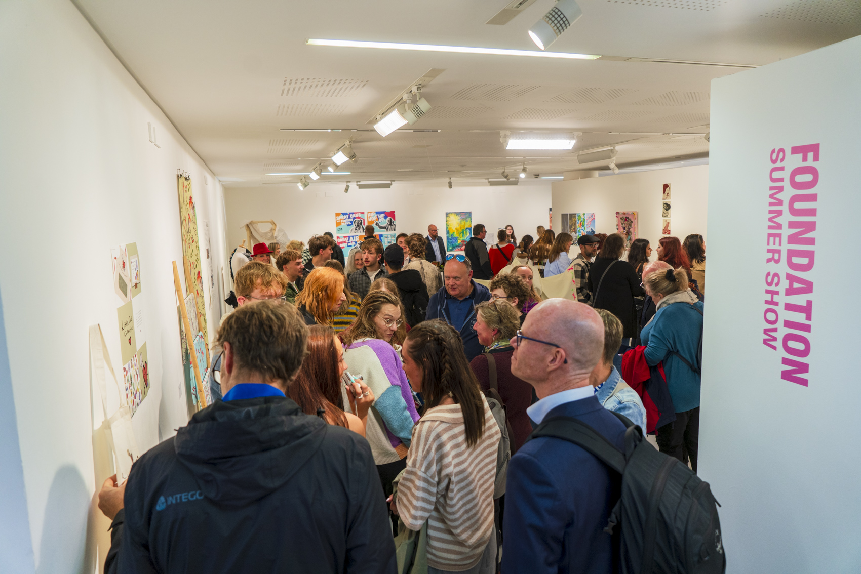 A crowded art gallery exhibition with visitors gathered around the room, under ceiling track lights and beside a wall marked 'FOUNDATION SUMMER SHOW'.