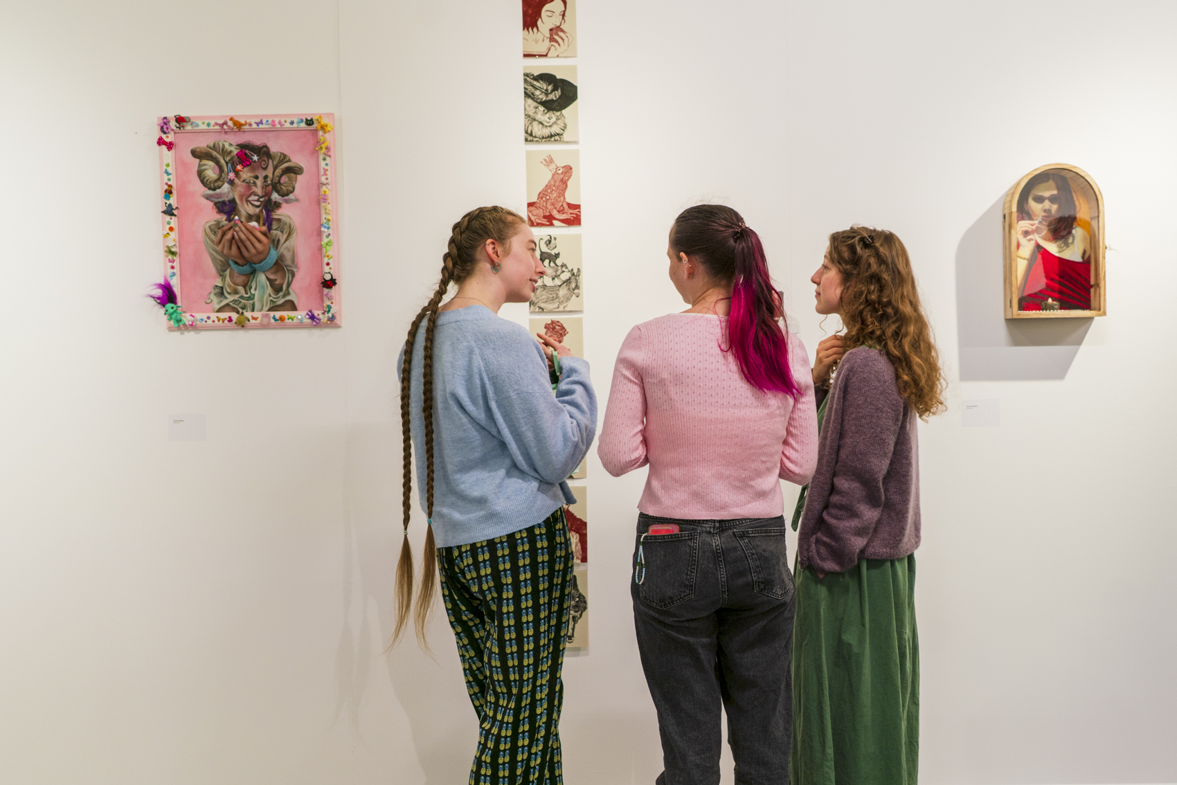Three people stand viewing a vertical art installation in a gallery, with framed artworks hung on the white walls.