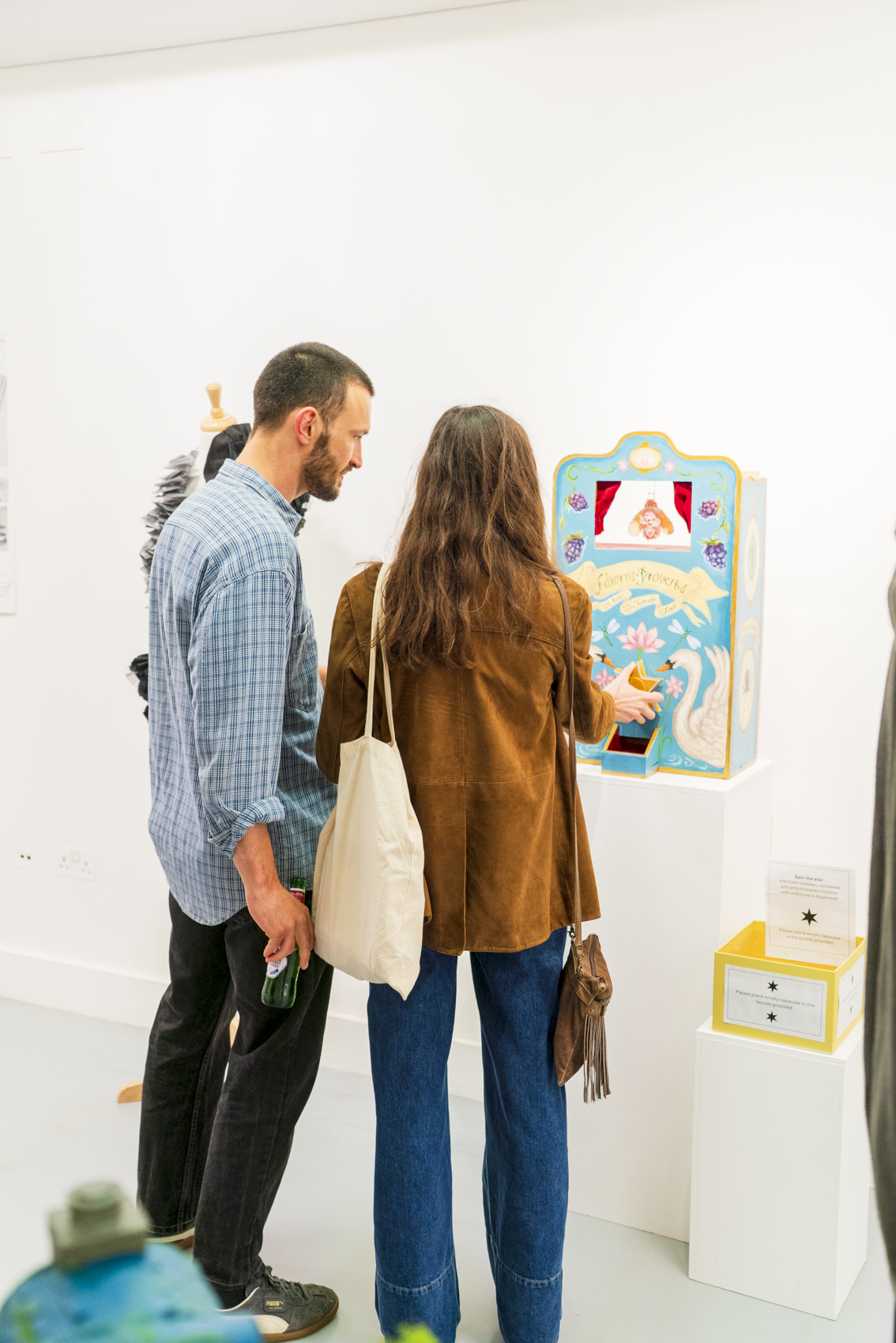Two people stand beside a colourful interactive display mounted on a white gallery wall, with other small display boxes on pedestals nearby.