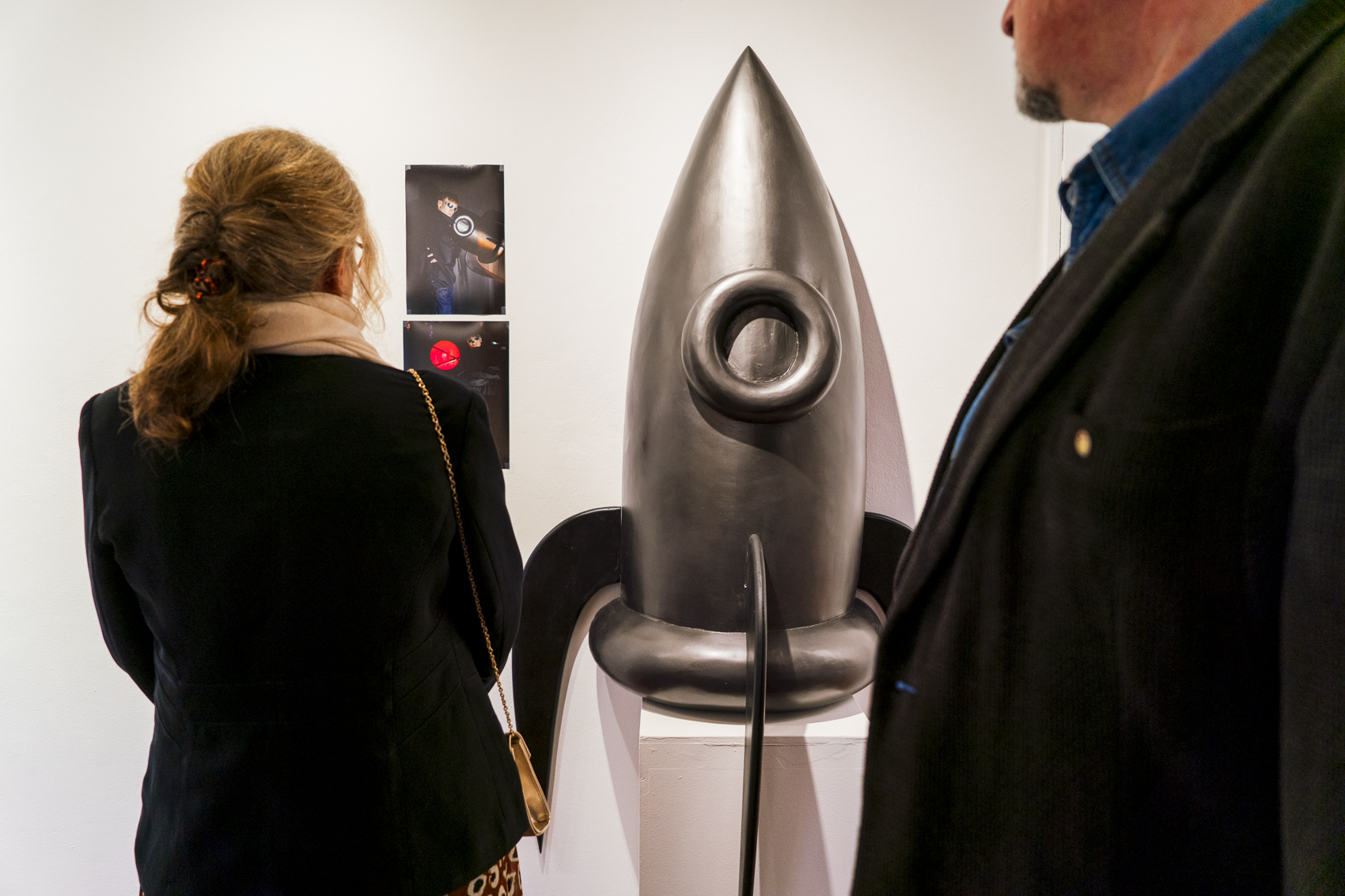 A metallic rocket sculpture stands on a white pedestal in a gallery, with two visitors viewing it from the foreground.