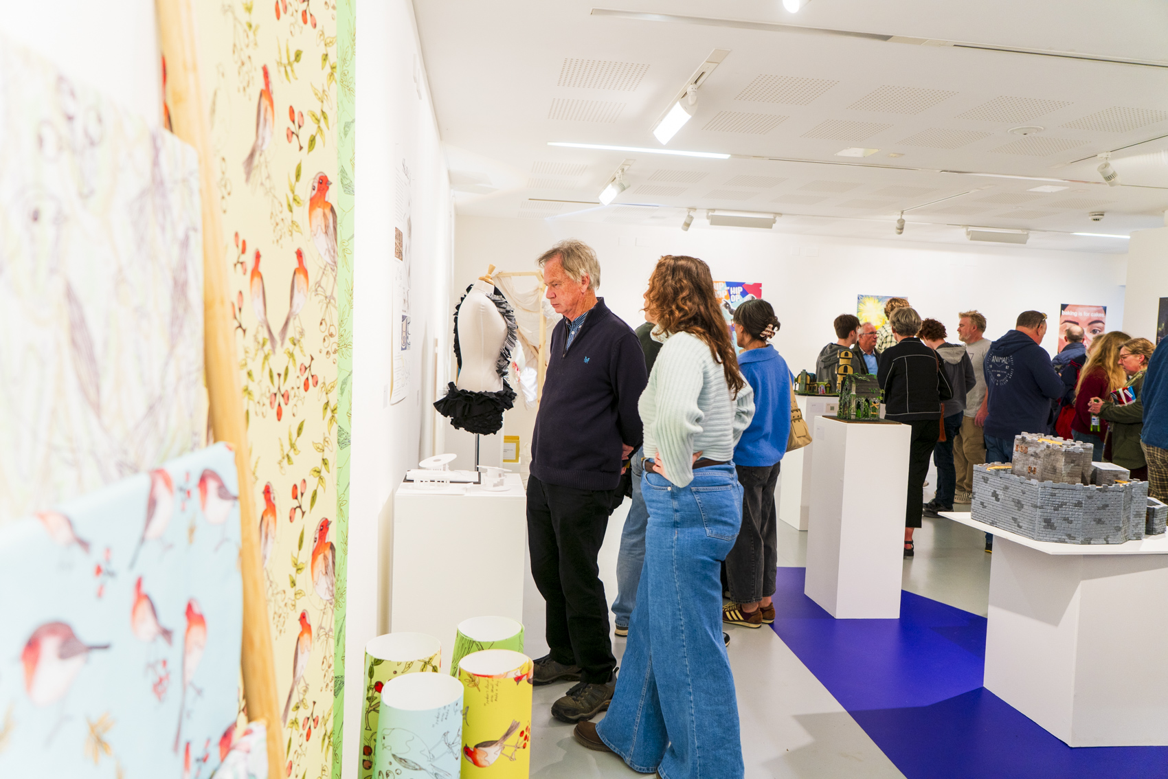 Visitors view textile and fashion displays in a bright gallery, with white plinths and patterned wall panels along a blue-and-white floor.