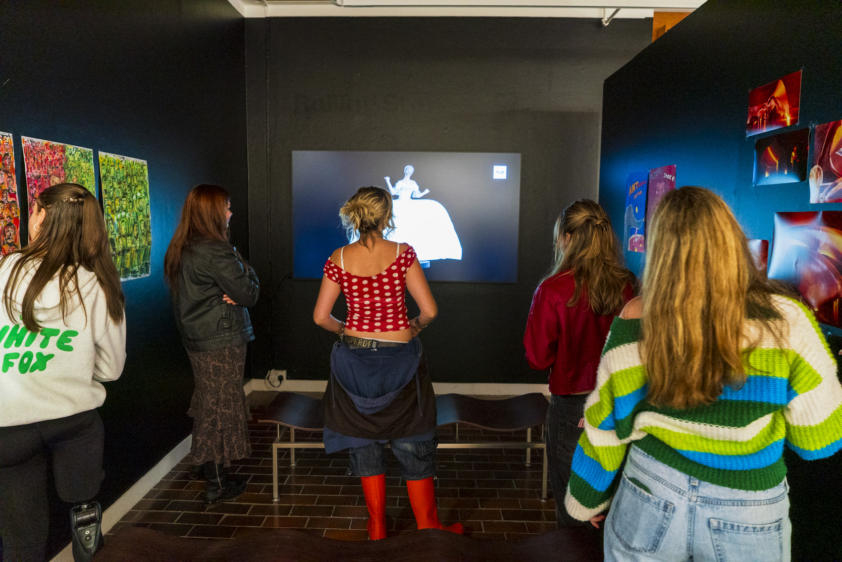 Five people stand in a dark gallery watching a screen, with framed artwork on the walls and a brick floor beneath them.