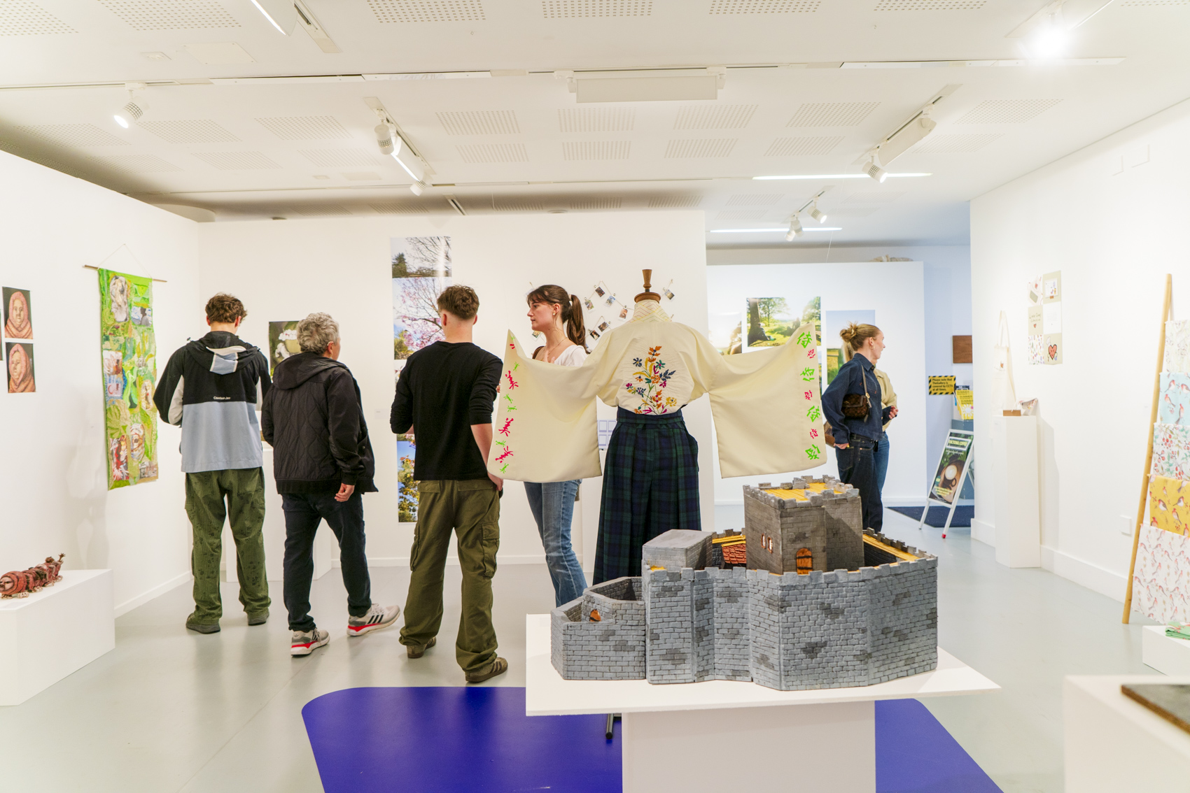 Several people view artworks in a bright gallery, with a mannequin displaying a floral embroidered jacket beside a table of grey woven baskets.