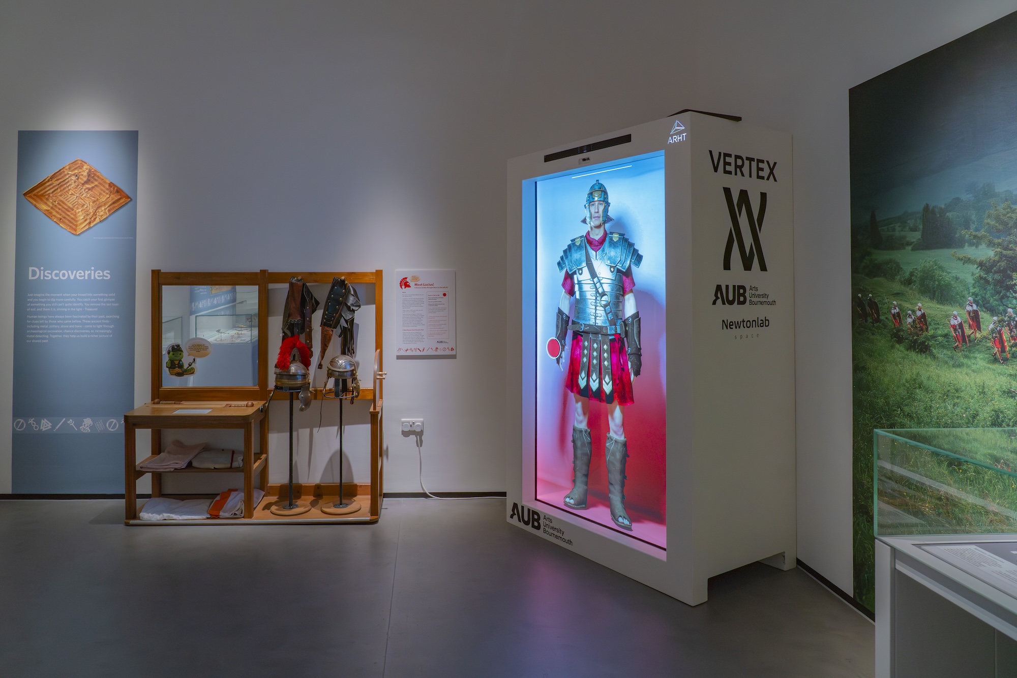 A museum exhibit showing Roman armour and helmets, next to a large white Holobox with a Roman soldier in full armour on the screen.