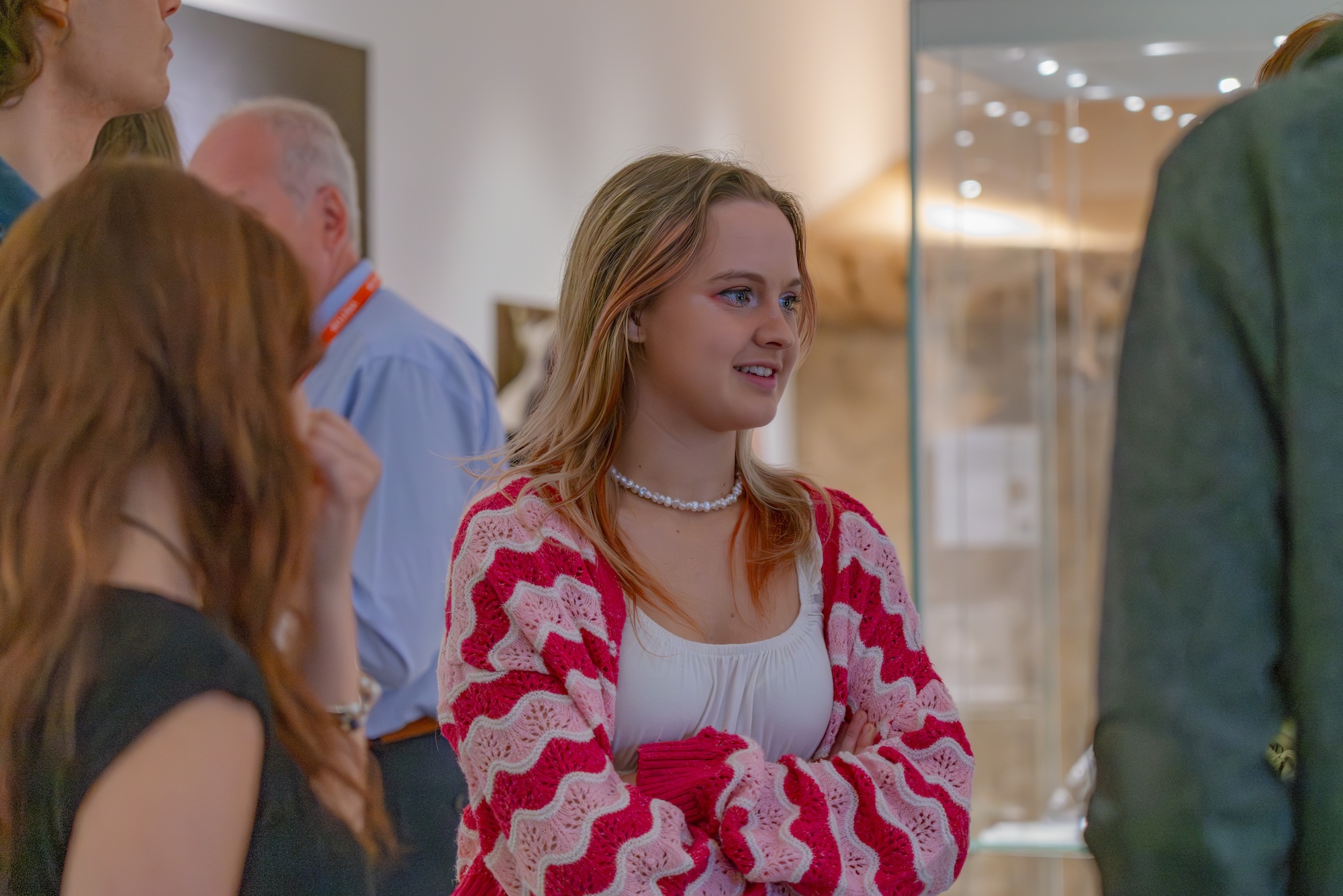 An Arts University Bournemouth student wearing a red and pink cardigan, arms crossed, while attending an exhibition at the Dorset Museum and Art Gallery.