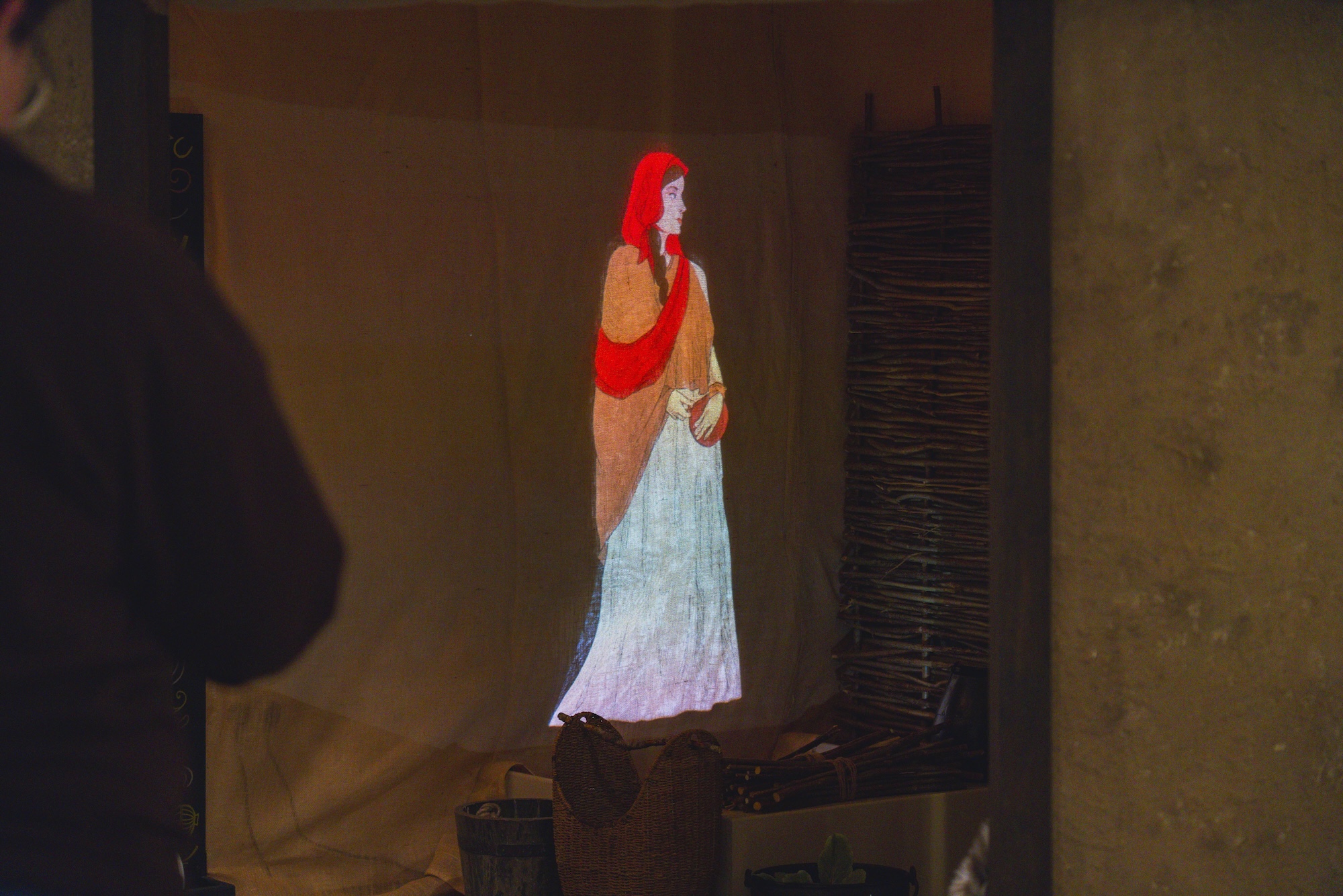 Projected image of a woman wearing a red head scarf, orange shawl and white robe, projected onto a canvas wall.