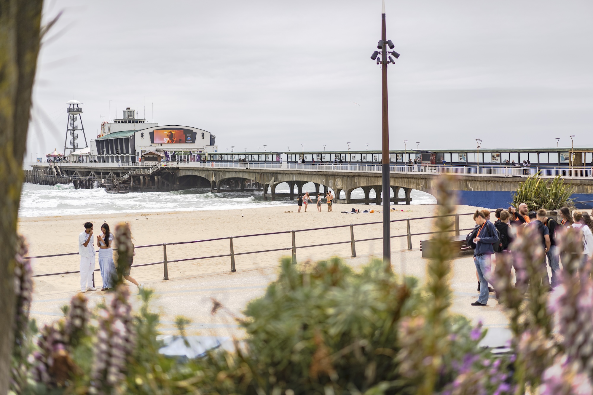 Bournemouth Pier with Bournemouth Beach and people walking on Bournemouth Pier Approach in the foreground on a cloudy day.