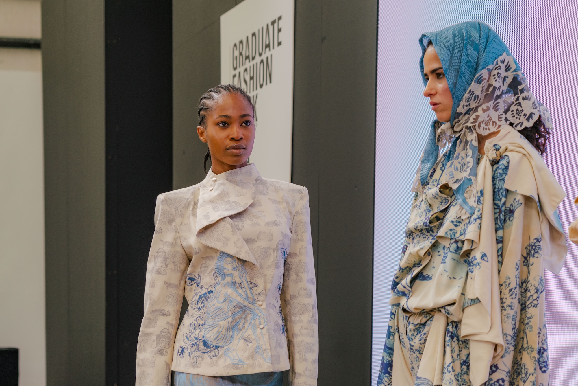 A model wearing a textured grey jacket with a high collar stands next to another model dressed in a patterned outfit featuring blue tones and a matching headscarf. The background displays a sign for "Graduate Fashion."