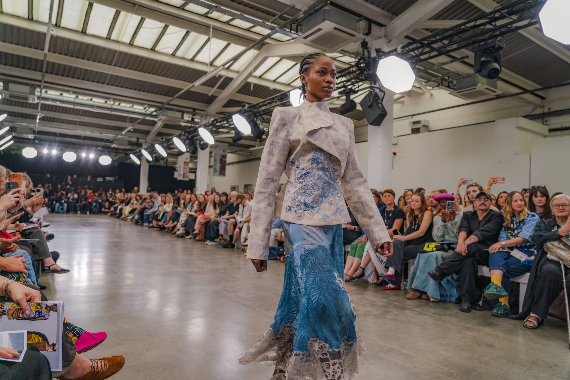 A model walks the catwalk in a patterned jacket and blue skirt, with an audience seated along both sides in an industrial fashion show venue.