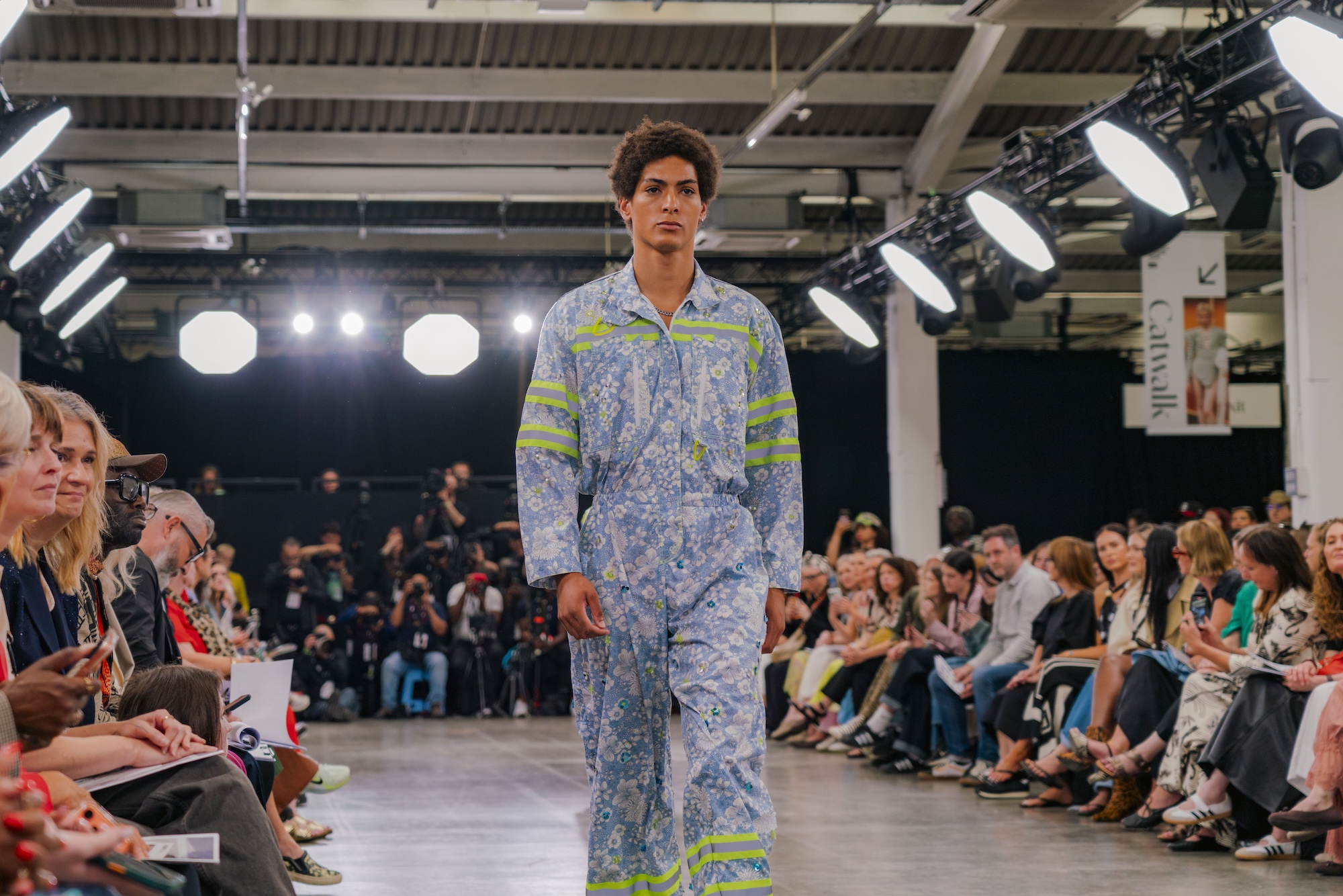 A model wearing a blue floral jumpsuit with green stripes walks down a runway during a fashion show. The audience on either side includes various individuals seated and watching the presentation.