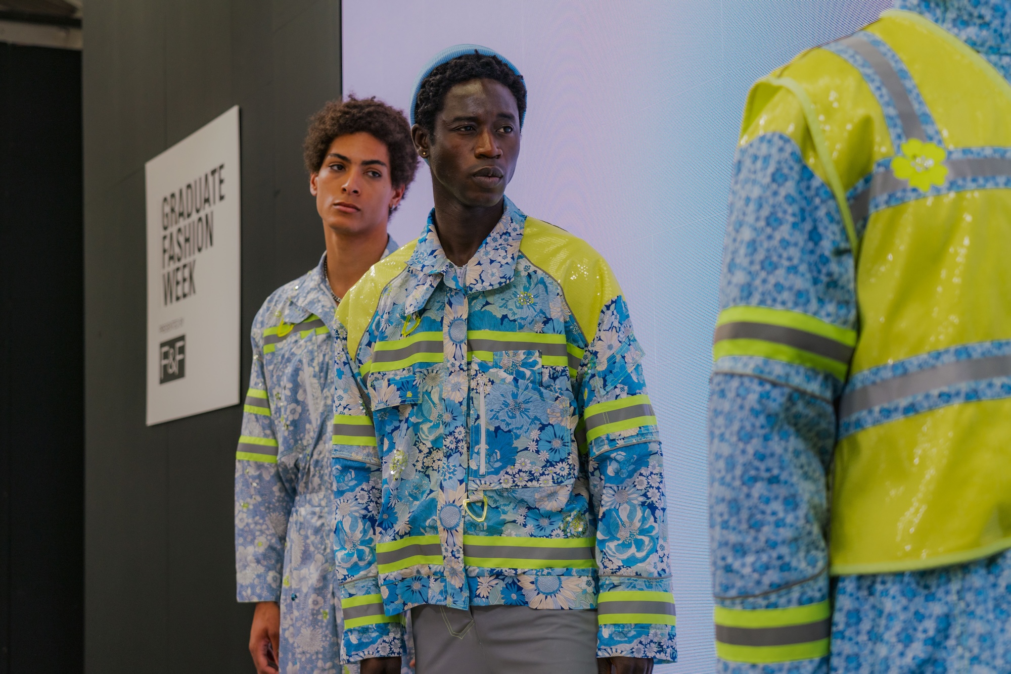 Two male models stand on a runway during a fashion event, one wearing a floral patterned jacket with reflective yellow accents and the other in a matching floral outfit. A backdrop with the text "Graduate Fashion Week" is visible.