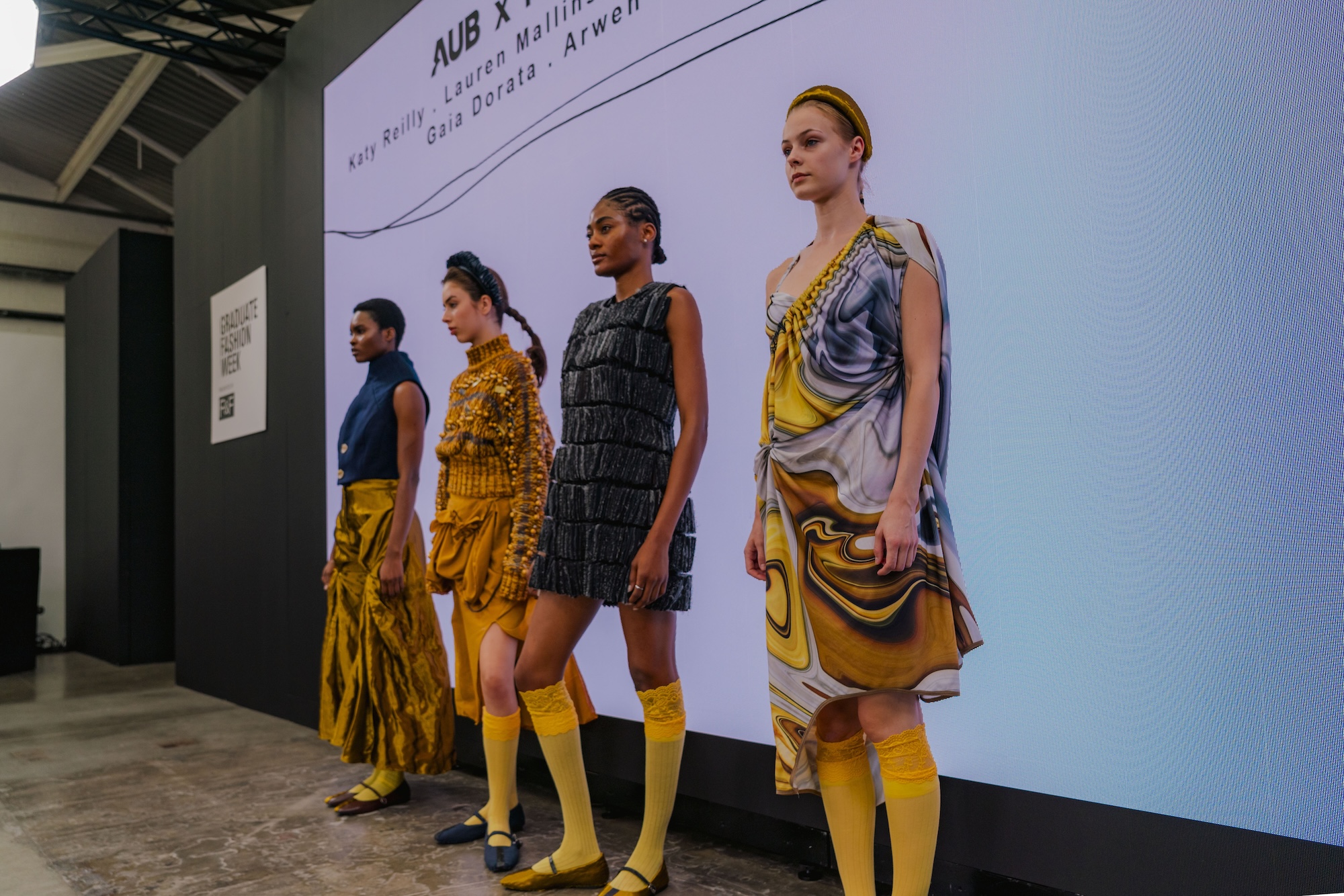 Four models are displayed wearing unique fashion designs in a runway setting. Each model showcases distinct outfits with various textures and colours, including yellow, blue, and intricate patterns, while standing against a large screen featuring event details.