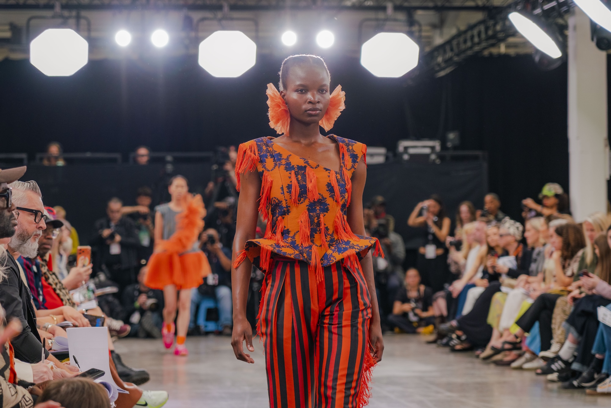 A model walks down the runway wearing a vibrant orange and blue patterned outfit with vertical stripes. The audience, seated on either side, watches, with some individuals holding cameras and phones to capture the moment.