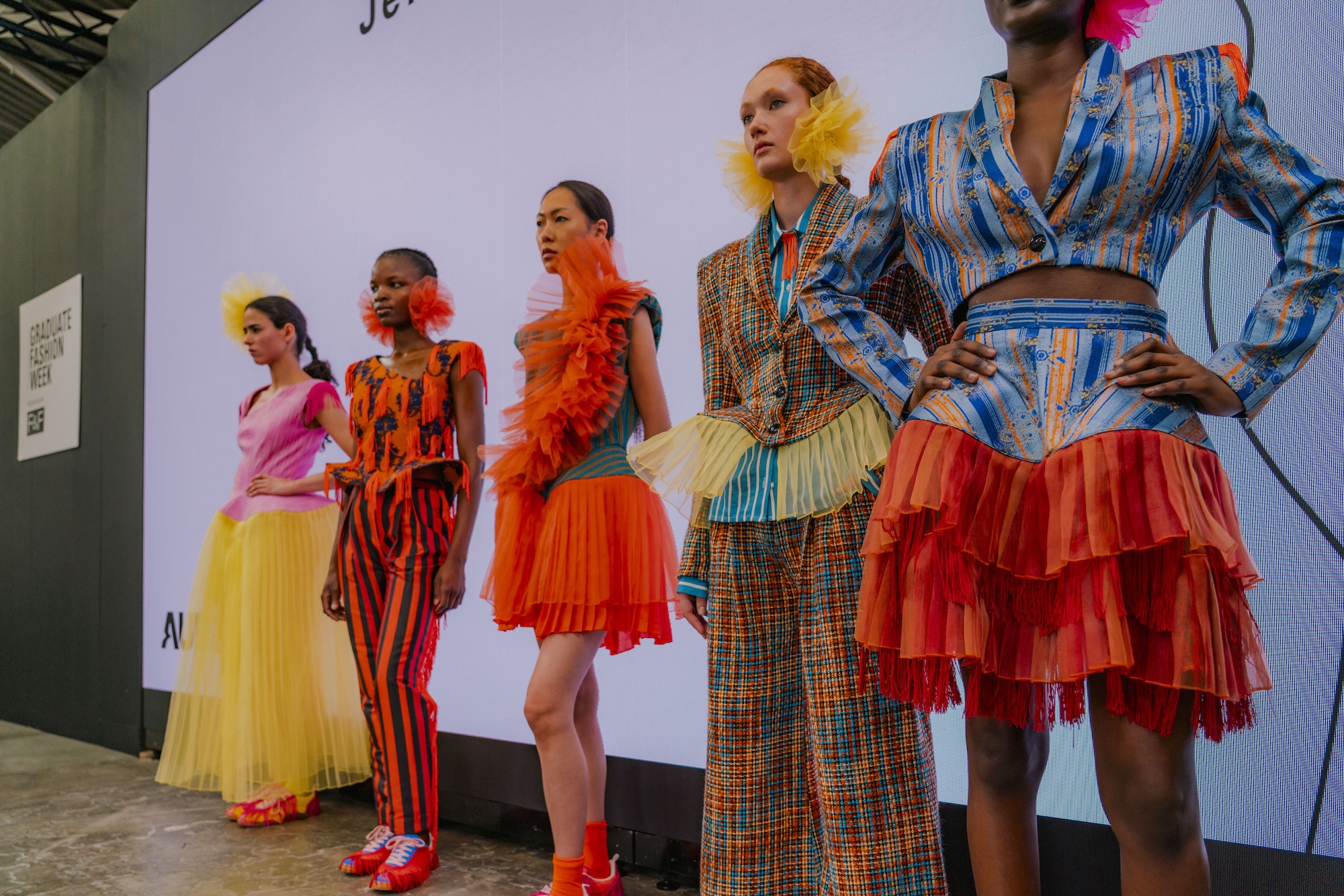 Five models showcase vibrant fashion designs on a runway. The first model wears a pink top and yellow skirt, followed by a model in orange with feather details. The third model features a striped suit, while the last two models exhibit colourful outfits with ruffles and bold patterns.
