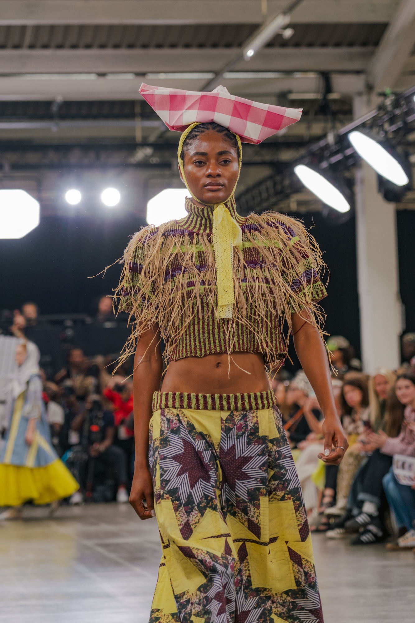 A model wearing a fringed top and patterned skirt walks down a runway at a fashion show, with seated spectators and bright studio lights in the background.