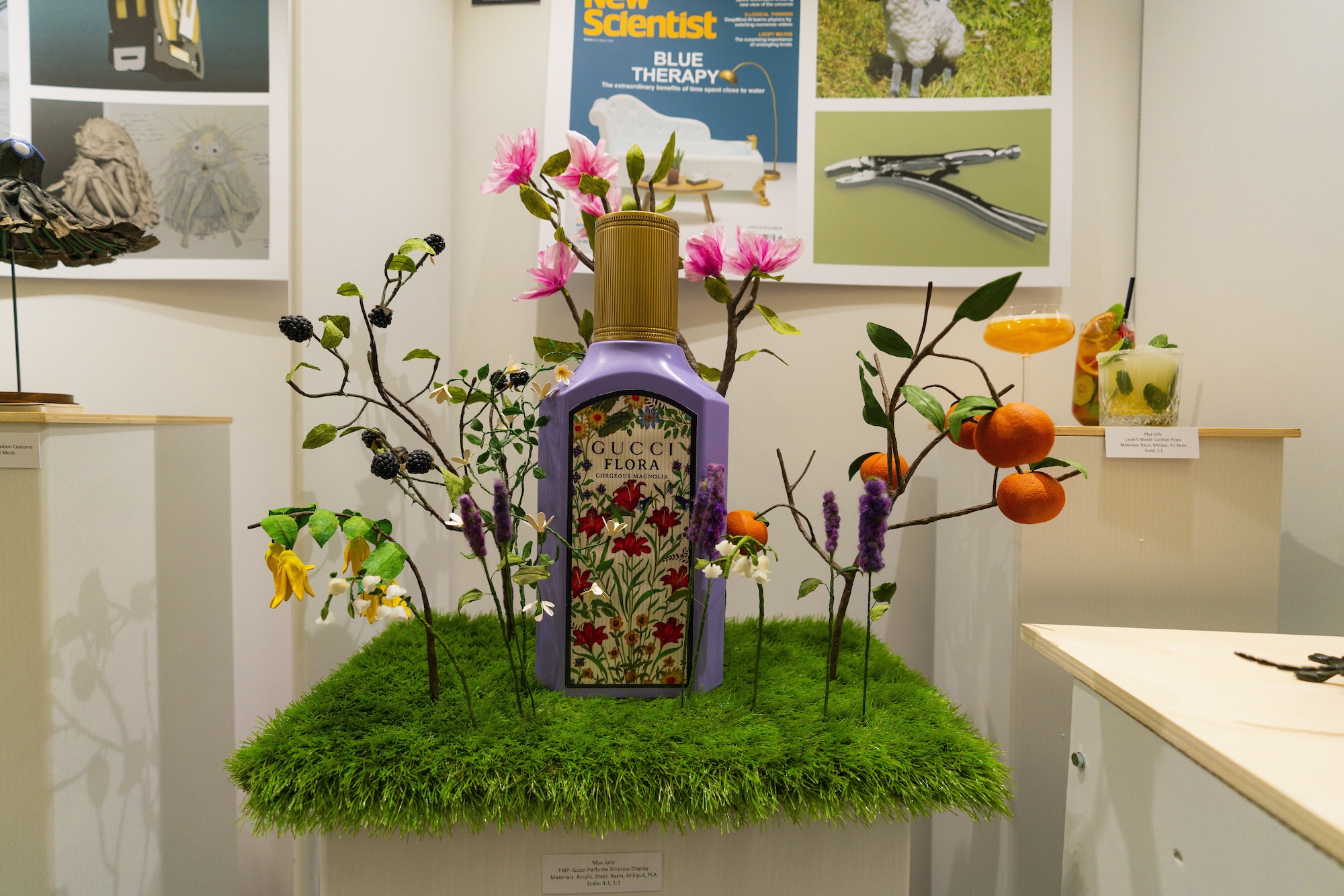 A decorative display featuring a bottle of Gucci Flora perfume surrounded by artificial flowers and fruits on a patch of green grass. The background includes various framed images and items mounted on a wall.