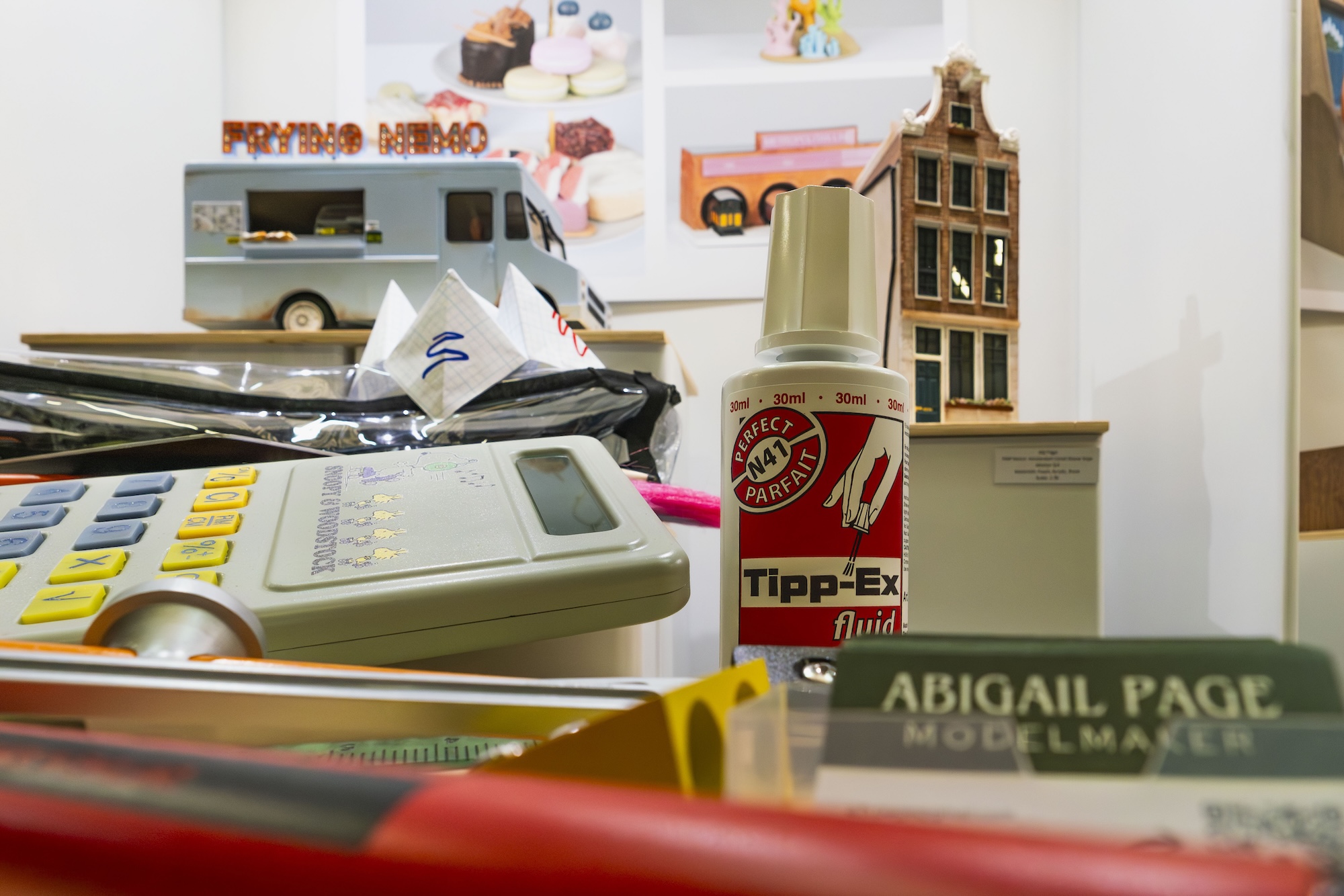 A collection of various items on a table includes a vintage calculator, a bottle of Tipp-Ex correction fluid, model making materials, and a small decorative building. In the background, there are images of cakes displayed.