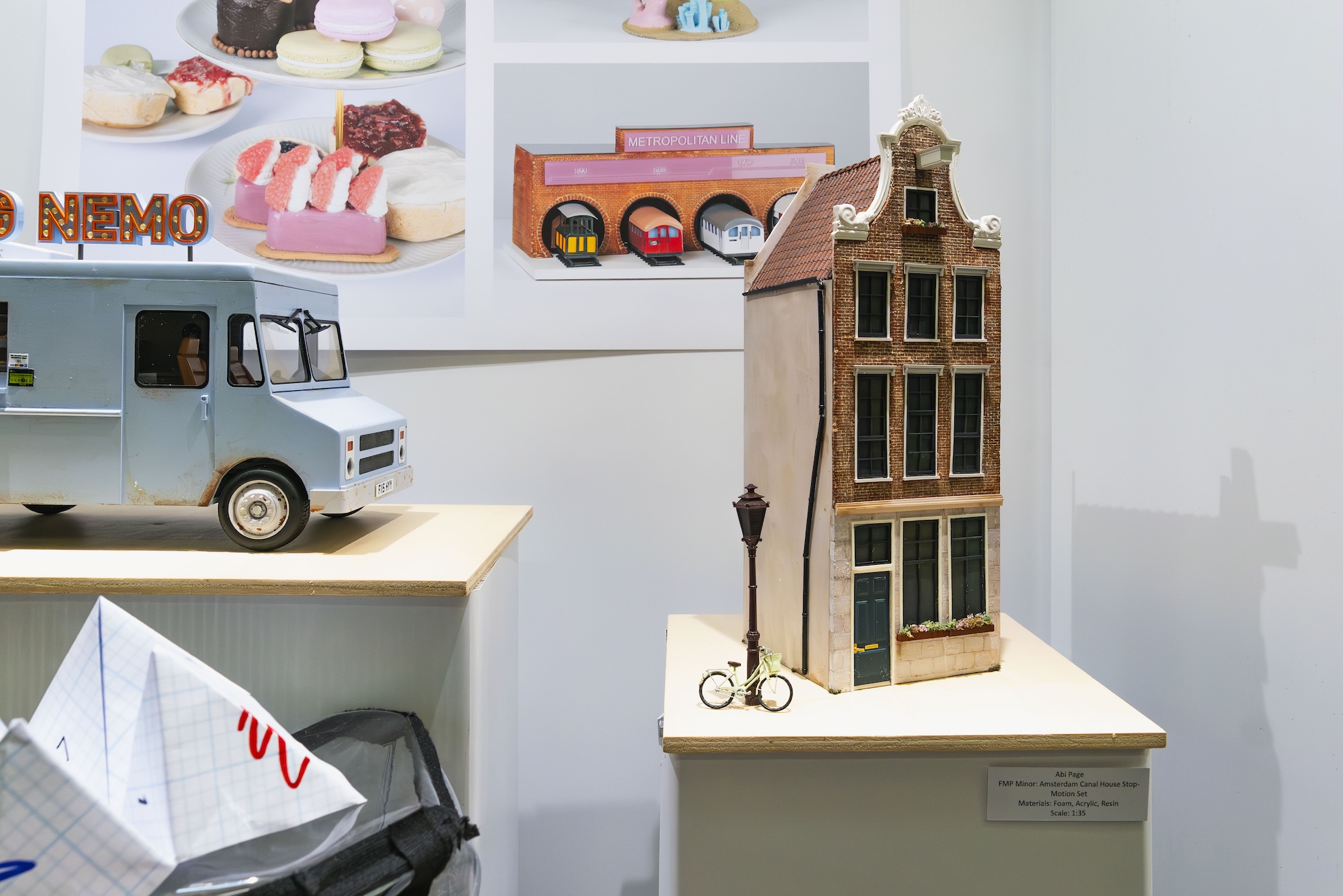 A decorative model of a narrow, brick building with large windows and a steep roof, displayed on a wooden pedestal. Beside it, a small bicycle is positioned next to a street lamp. In the background, a poster features various desserts.