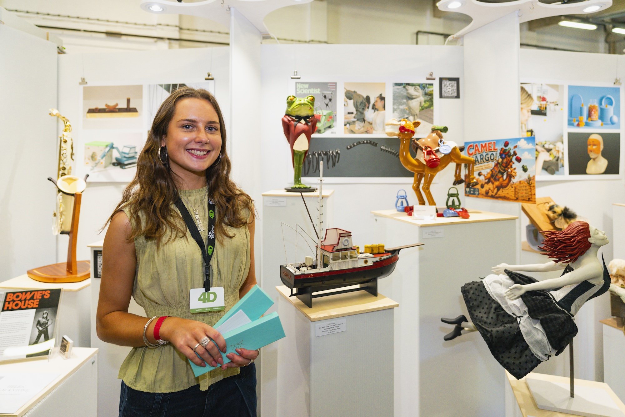 A young woman with long hair, wearing a beige top and a black lanyard, stands in an exhibition space. She holds two blue booklets and is positioned in front of various displayed art pieces and sculptures on white pedestals.
