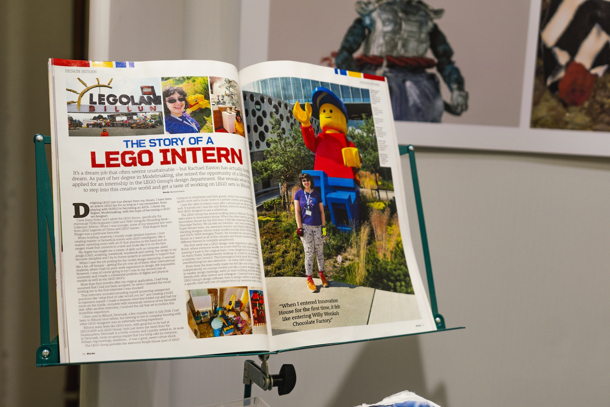 An open magazine on a stand displays an article titled "THE STORY OF A LEGO INTERN" alongside an image of a person standing next to a large LEGO figure. The background features a blurred view of a display with LEGO models.