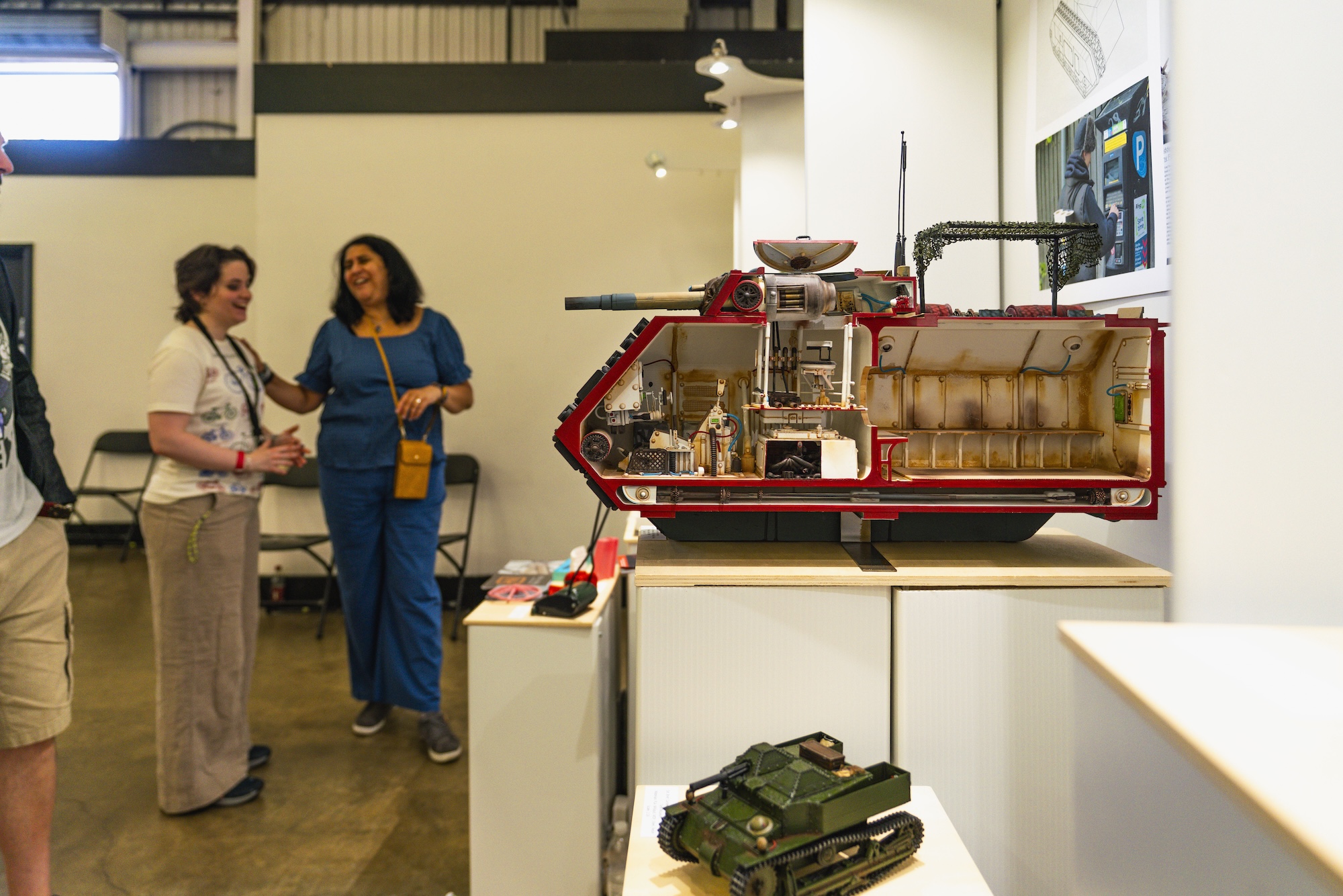 A detailed model of a vehicle displayed on a wooden shelf, showing its interior components. Two women stand nearby, engaged in conversation, while a small tank model sits on the shelf beside the larger vehicle model in a brightly lit exhibition space.