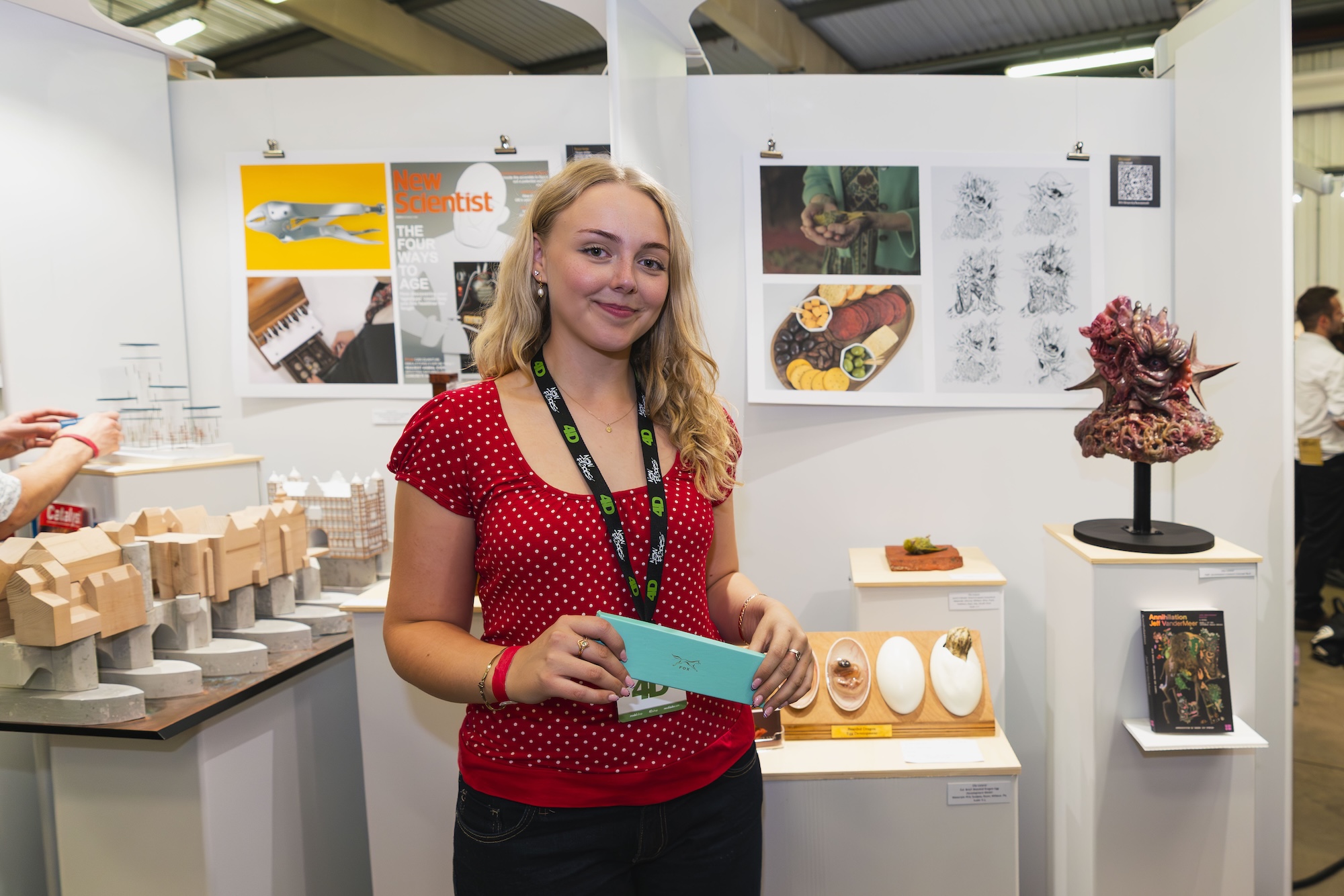 A woman with long blonde hair, wearing a red polka dot top, stands in front of an exhibition display. The display features various artworks, including printed images, sculptures, and a model made of wood.